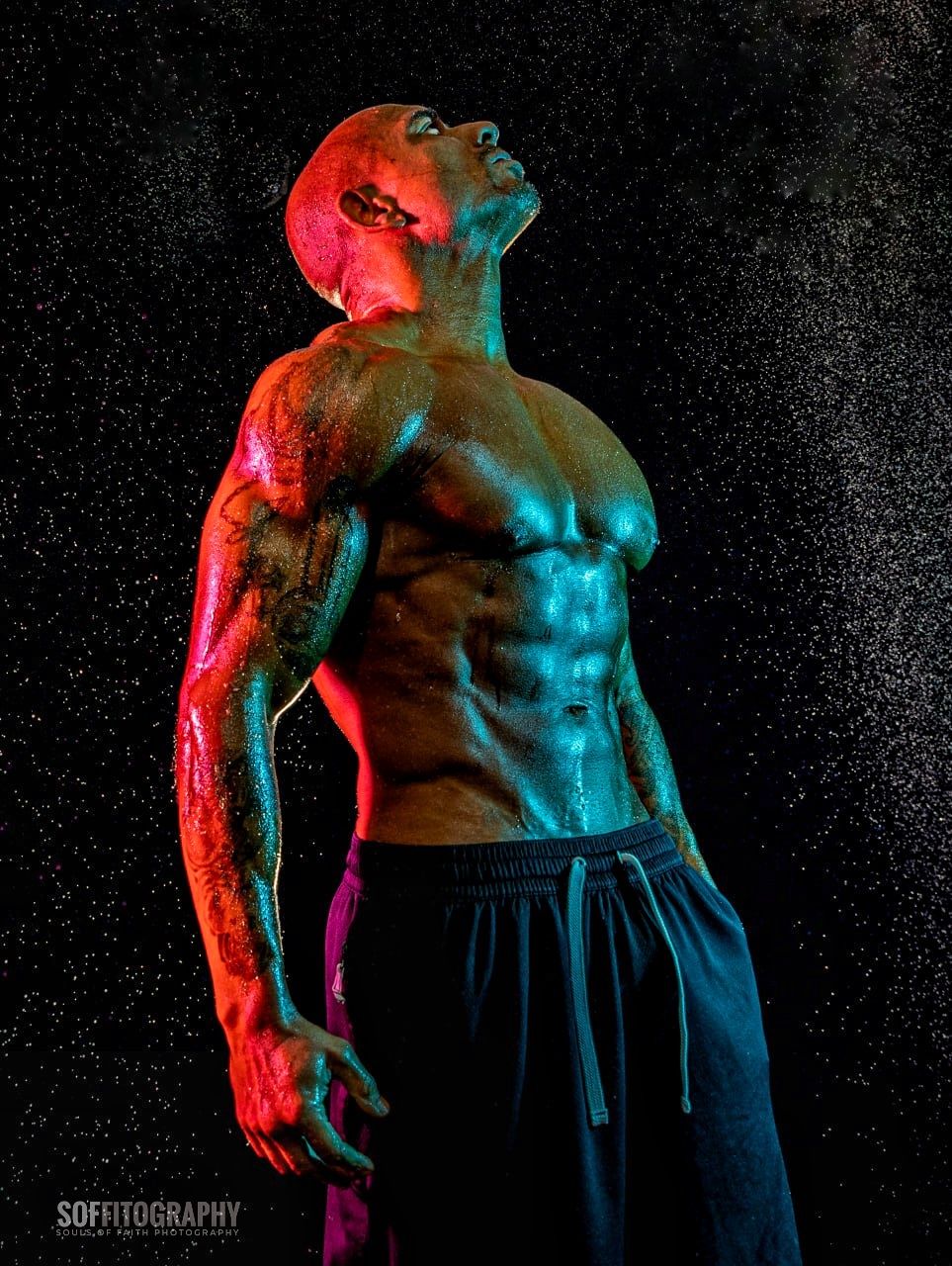 Muscular shirtless man with tattoos posed dramatically under colorful red and teal studio lighting with water droplets.