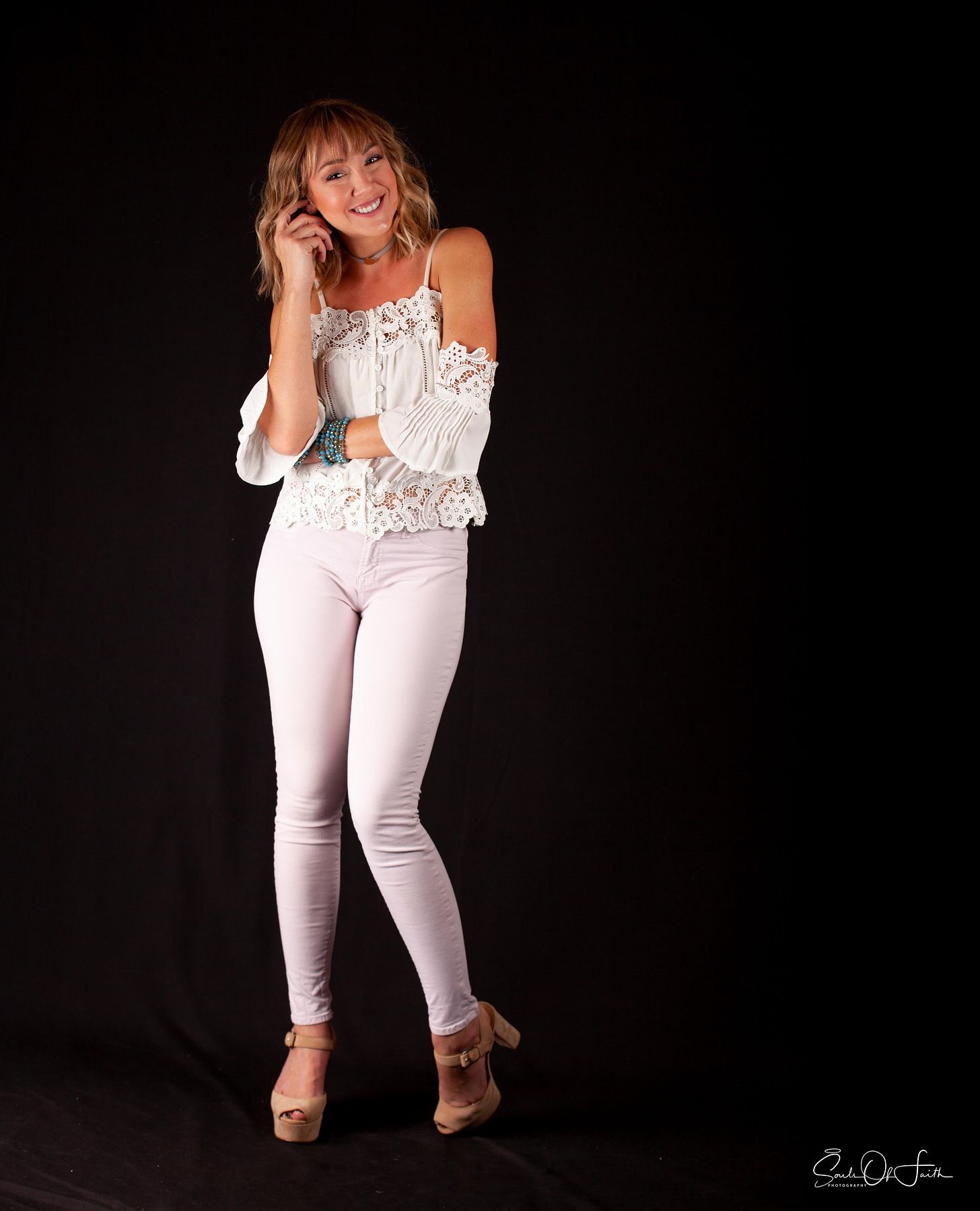 Young woman in white lace off-shoulder top and light pink skinny jeans posing confidently against black background.
