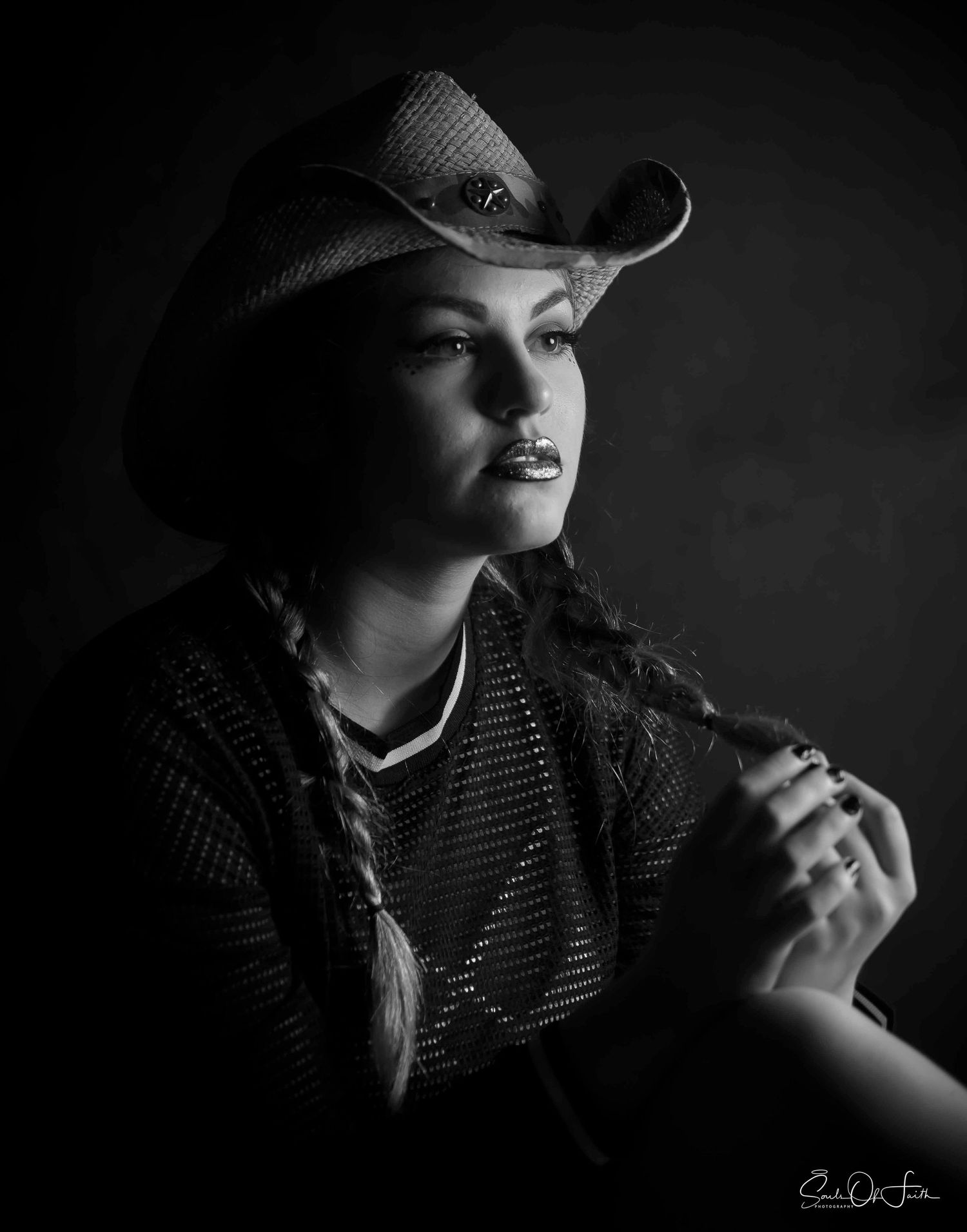 Black and white portrait of a young woman in a cowboy hat with braids, posing dramatically in studio lighting.