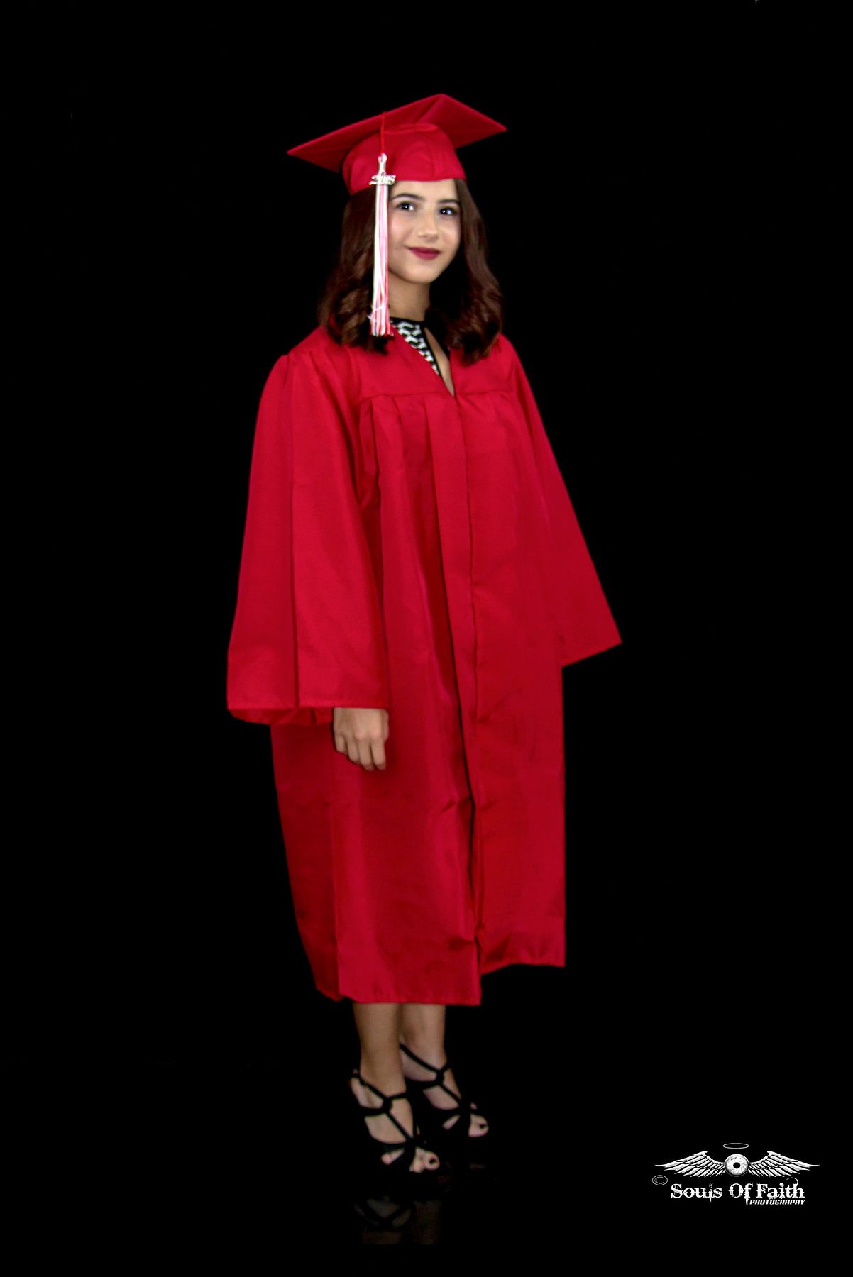 Young woman in red graduation cap and gown posing proudly against a black background for her graduation portrait.