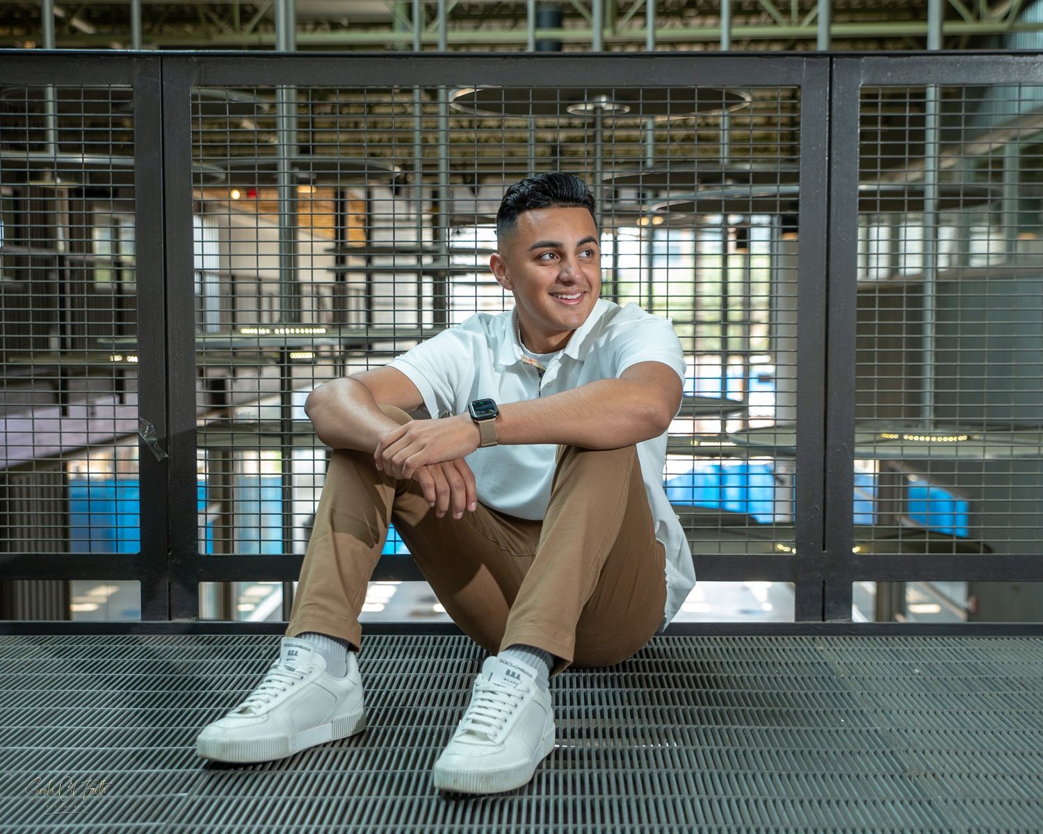 Young man in white polo and khaki pants sits smiling on metal grate floor in modern industrial space.