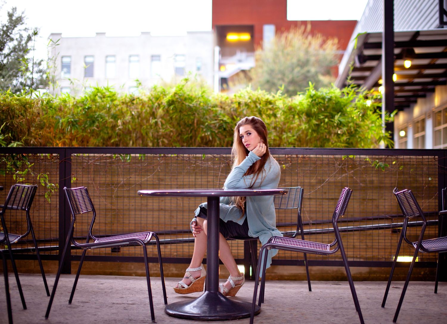 Young woman sitting alone at outdoor cafe patio table, surrounded by lush greenery and urban buildings.