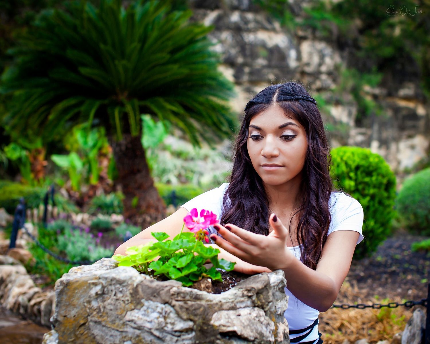 Young woman with long dark hair examines a pink flower in a lush garden with palm trees and stone walls.