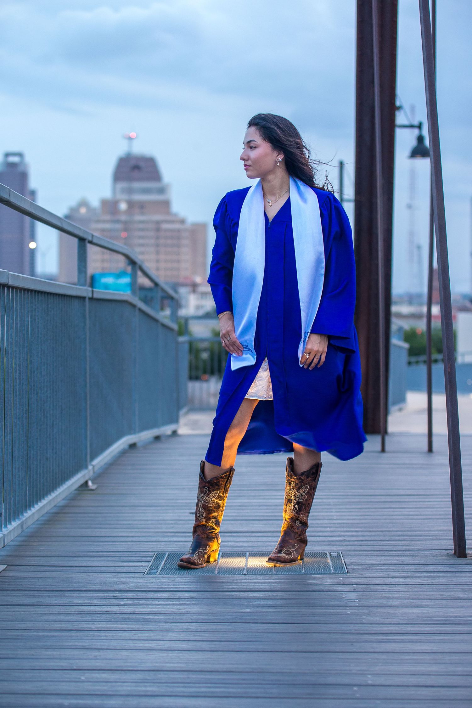 Woman in royal blue dress, white scarf, and cowboy boots stands on wooden boardwalk with city skyline at dusk.