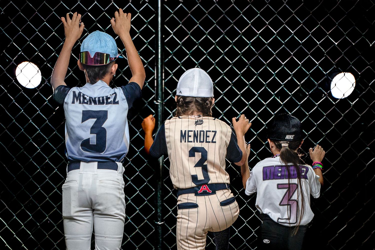 Three players named Mendez wearing jersey number 3 stand at a chain-link fence during a softball game.