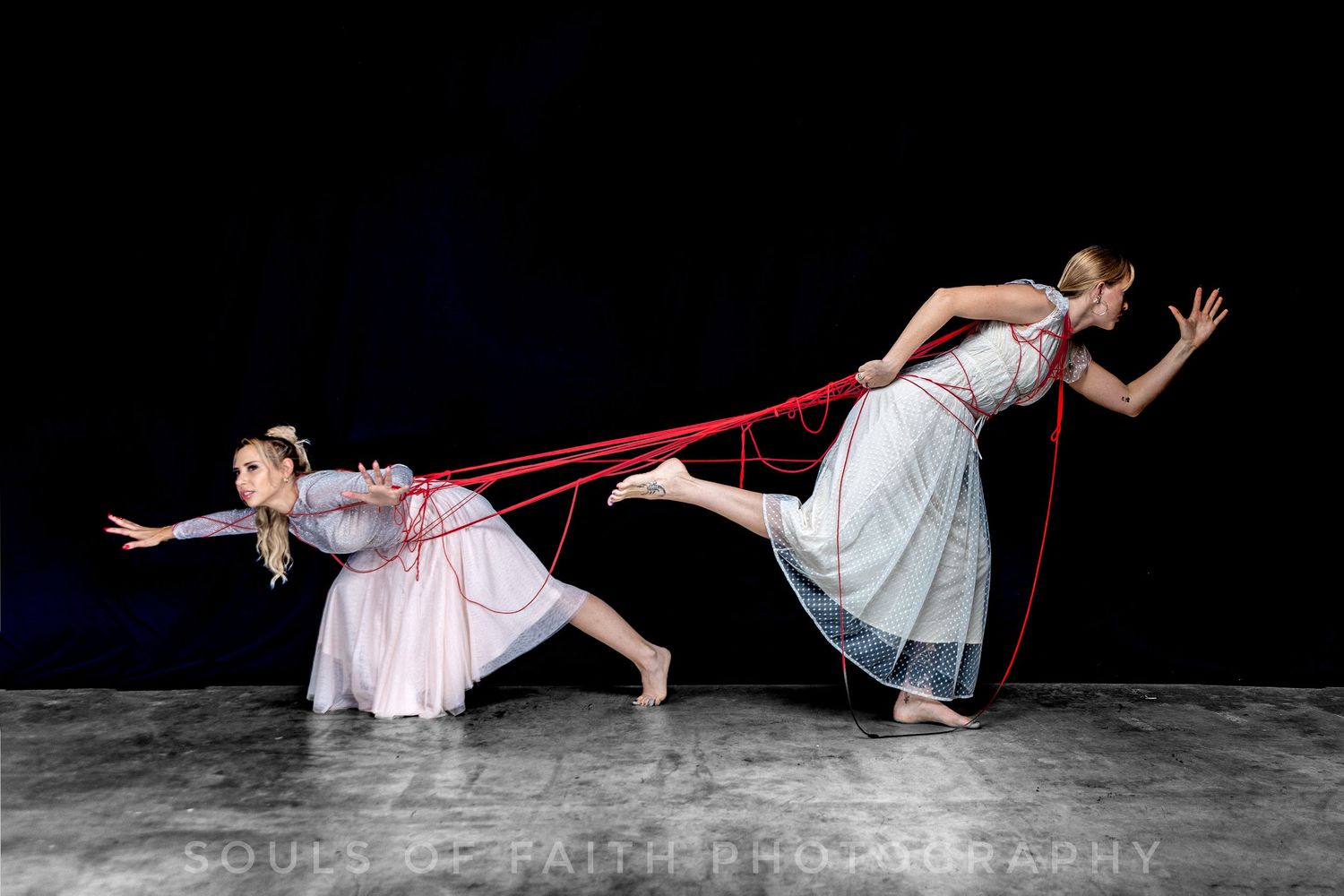 Two dancers in white dresses connected by red ribbons perform a dramatic contemporary dance on a dark stage.