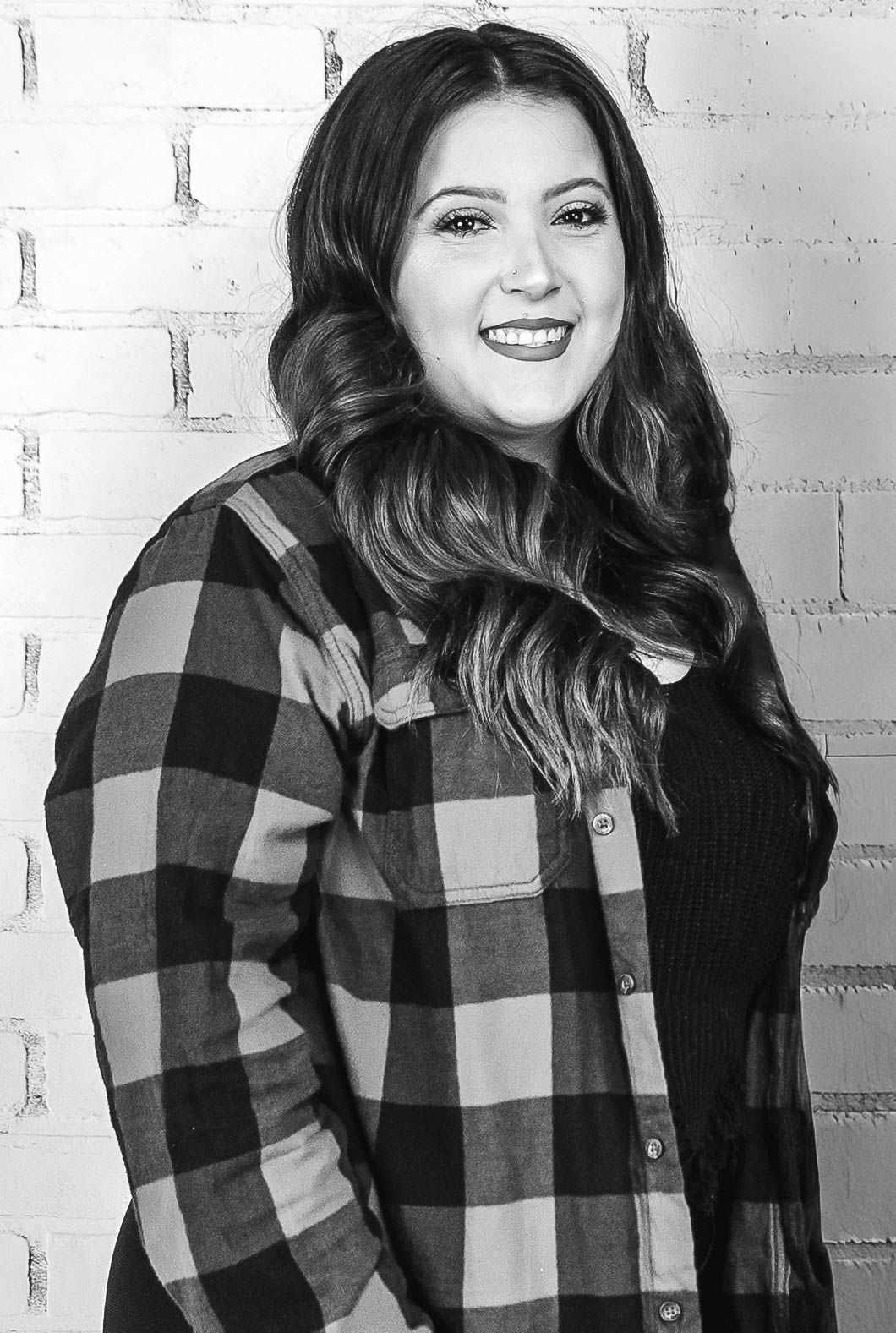 Smiling woman with long wavy hair wearing a plaid flannel shirt, posing against a brick wall in black and white.