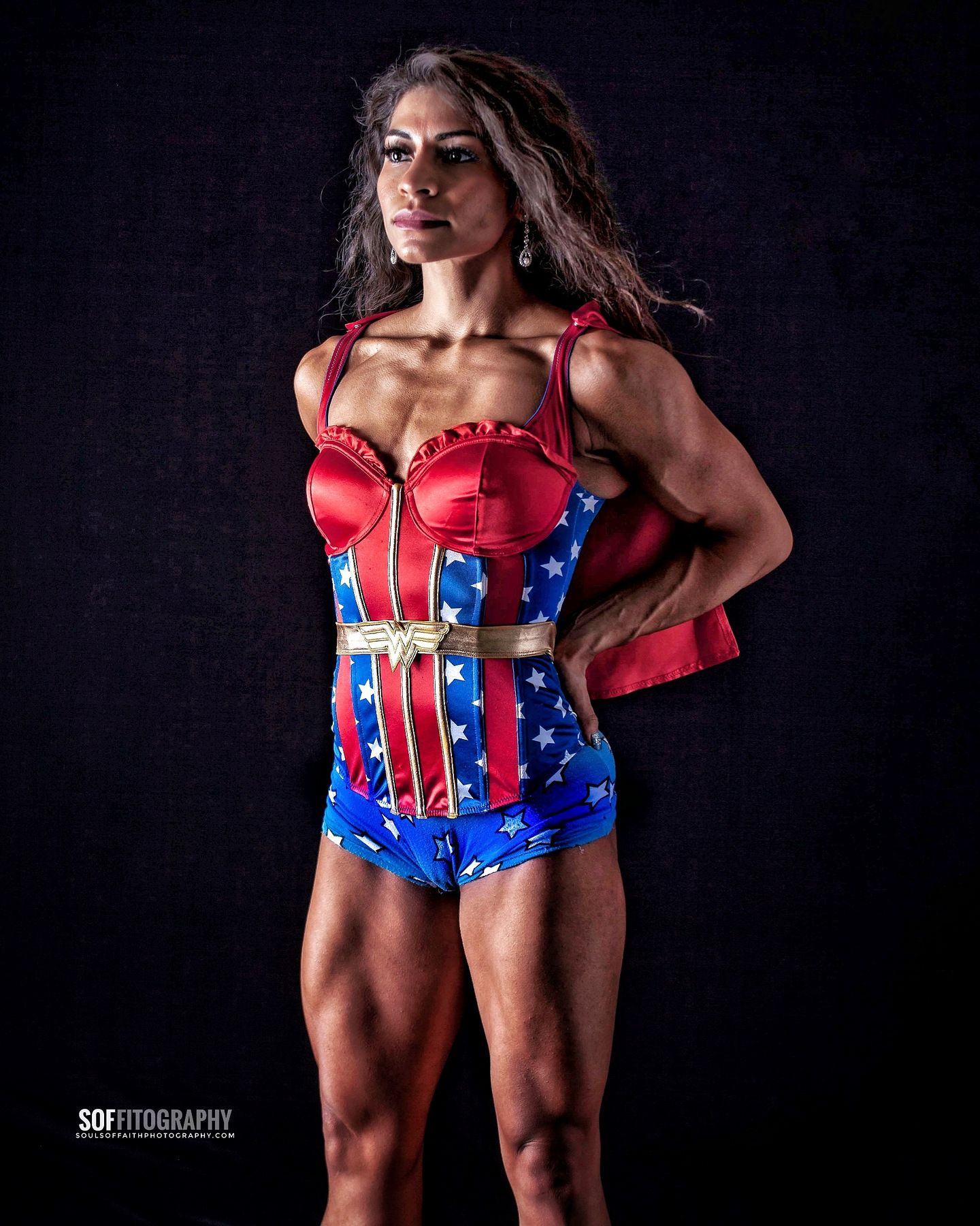 Muscular woman posing confidently in a Wonder Woman-inspired red, blue, and gold corset costume against a dark background.