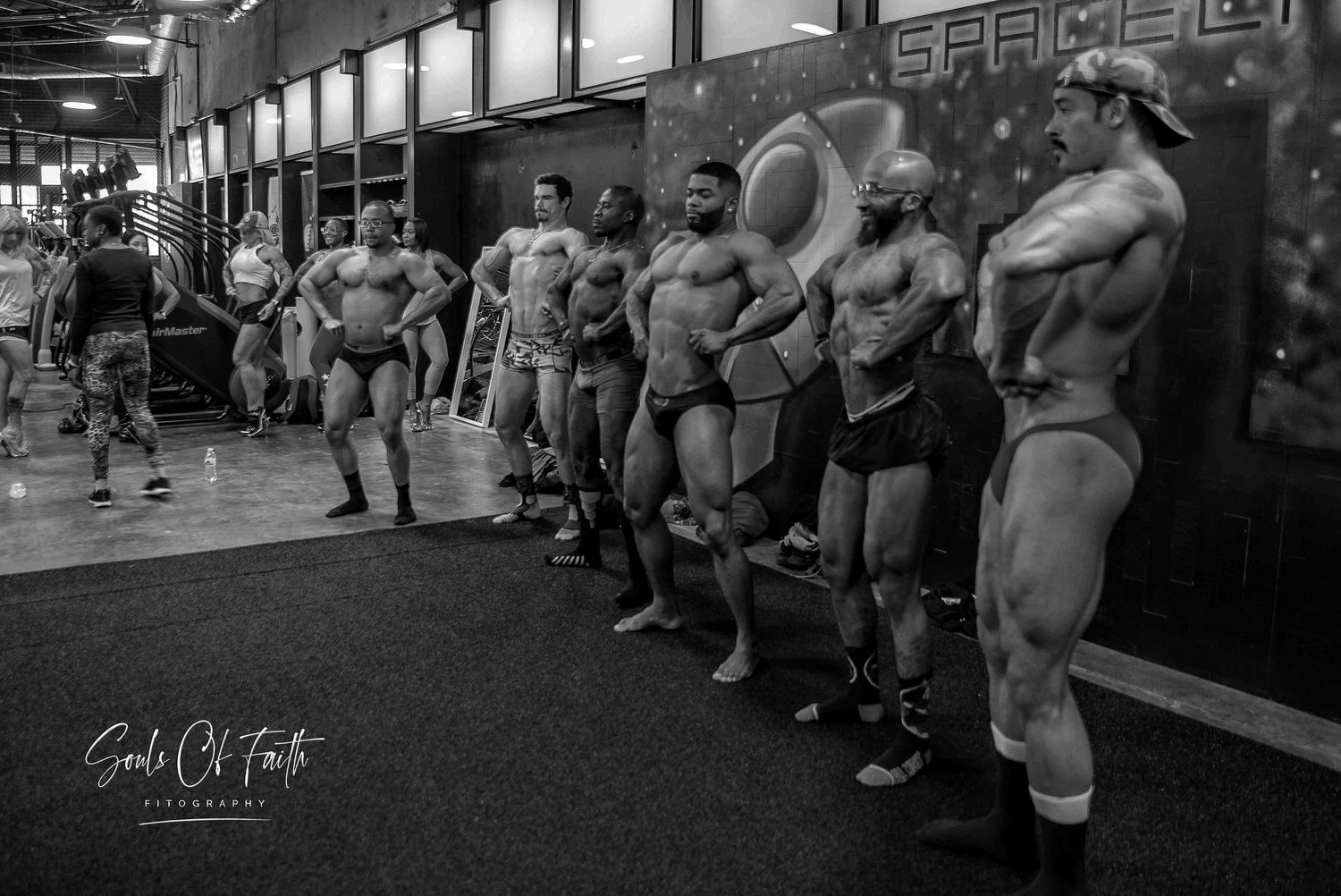Black and white photo of muscular bodybuilders lined up on stage at a fitness competition in a gym.