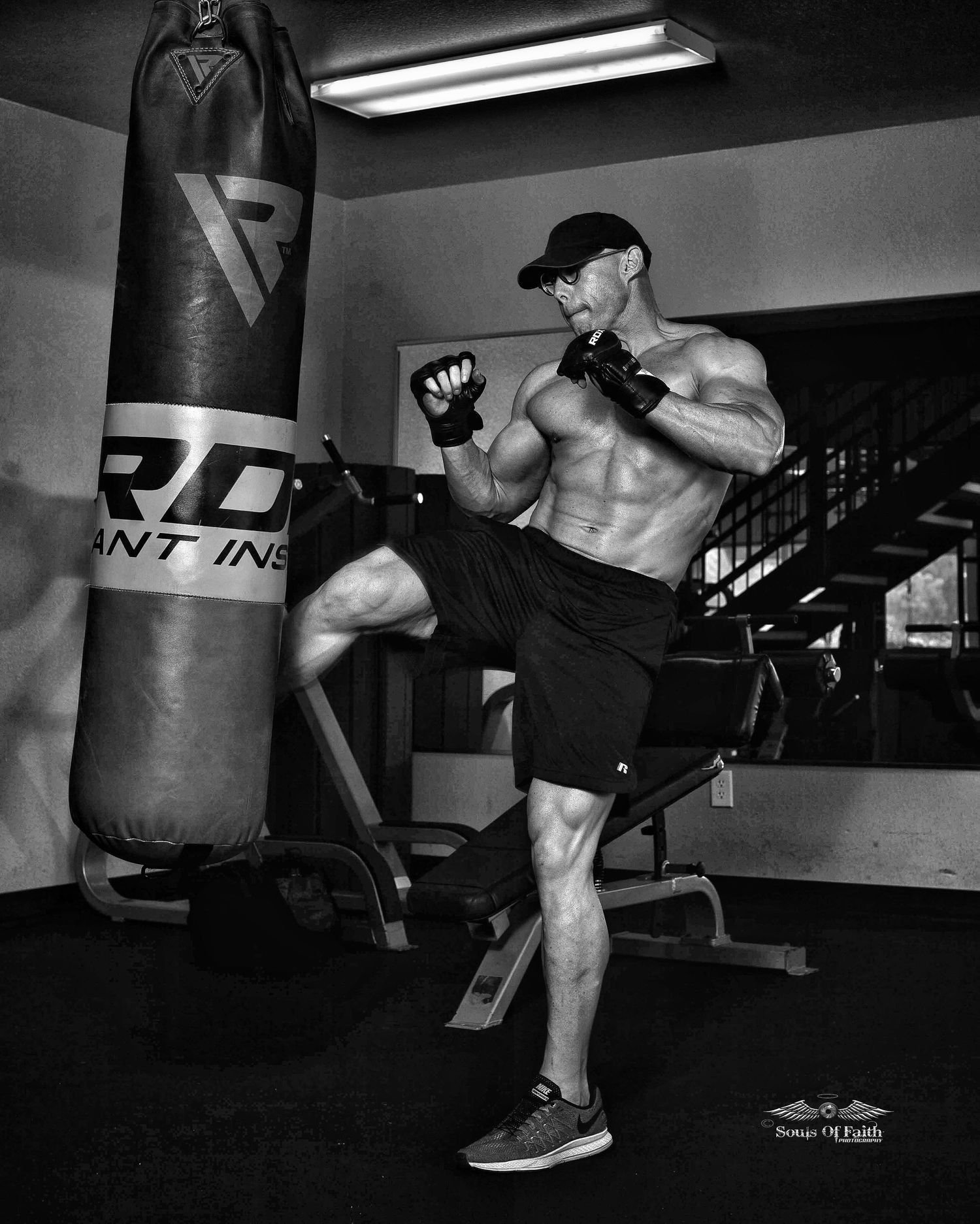 Muscular shirtless man performing a high kick at a heavy punching bag in a gym, black and white fitness photo.