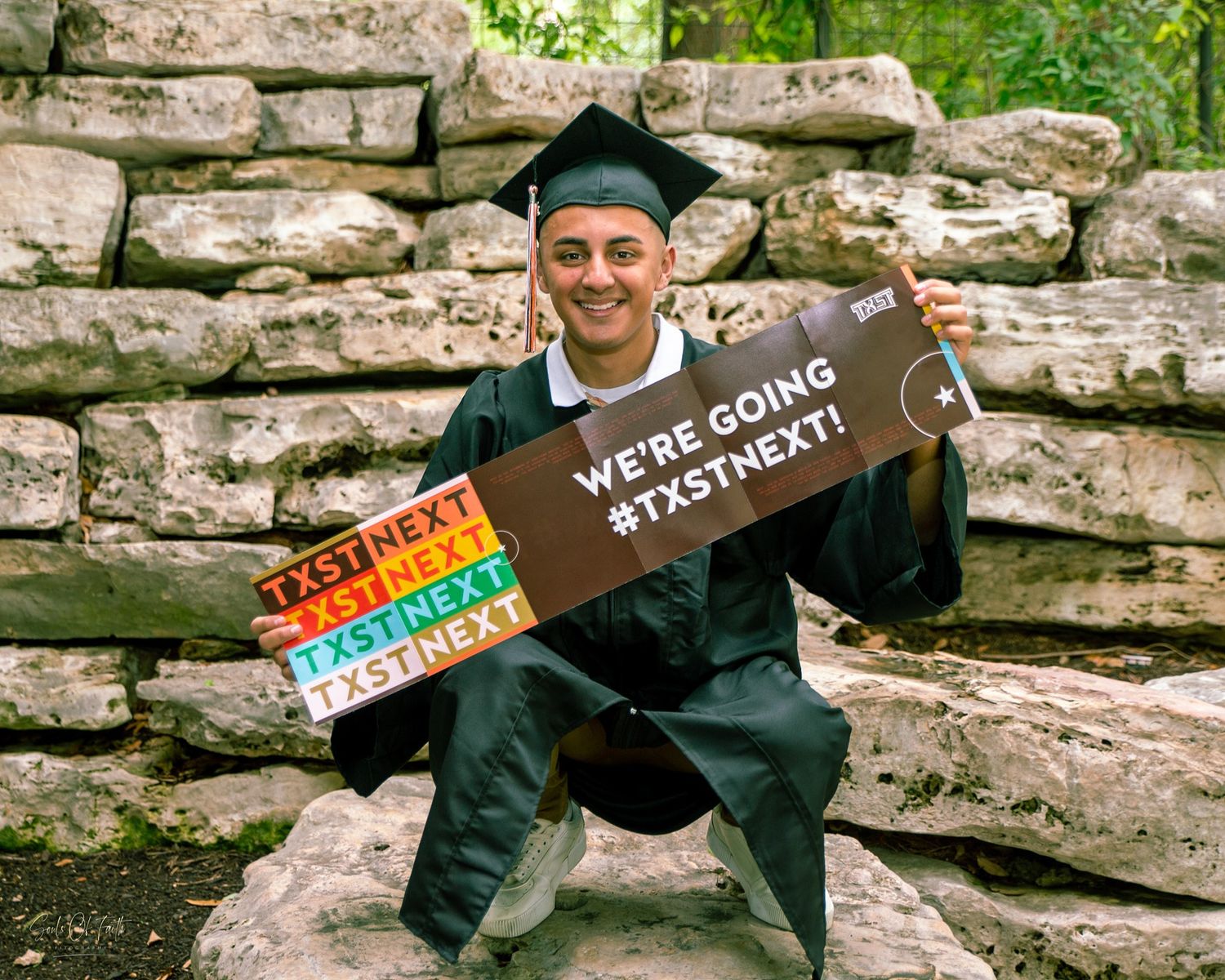 Graduate in cap and gown holding #TXSTNEXT signs, celebrating college acceptance outdoors near stone wall.