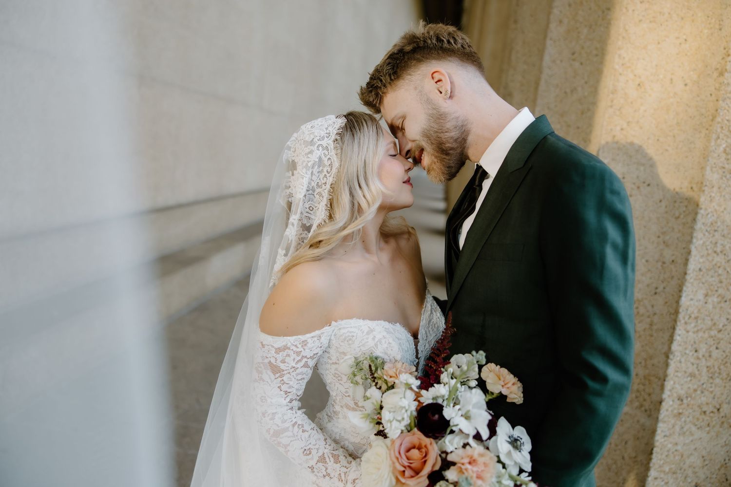 Bride and groom share a romantic moment, touching foreheads, bride in lace gown with veil holding floral bouquet.