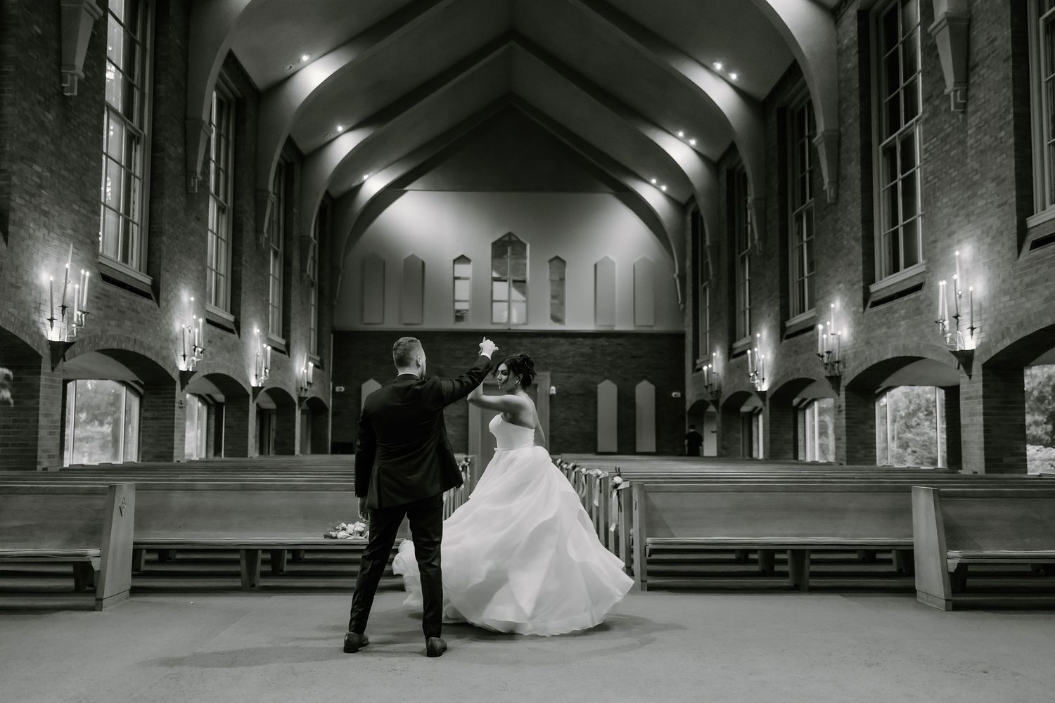 Black and white photo of newlyweds dancing in a dimly lit church hall with vaulted ceilings.