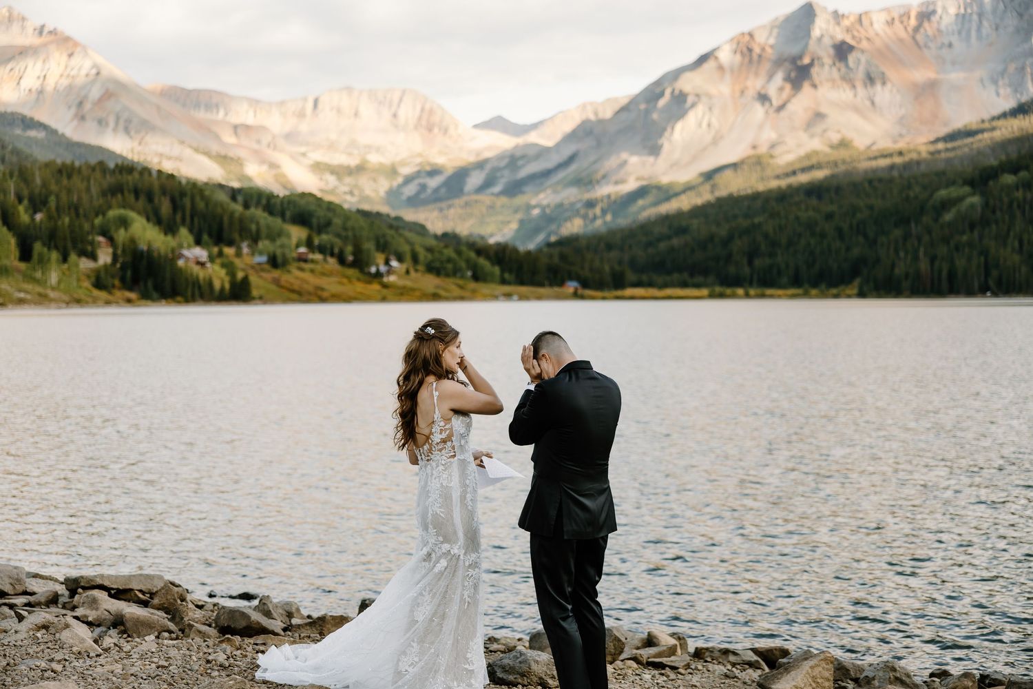 Panoramic mountain lake setting frames an intimate wedding ceremony on rocky shore.