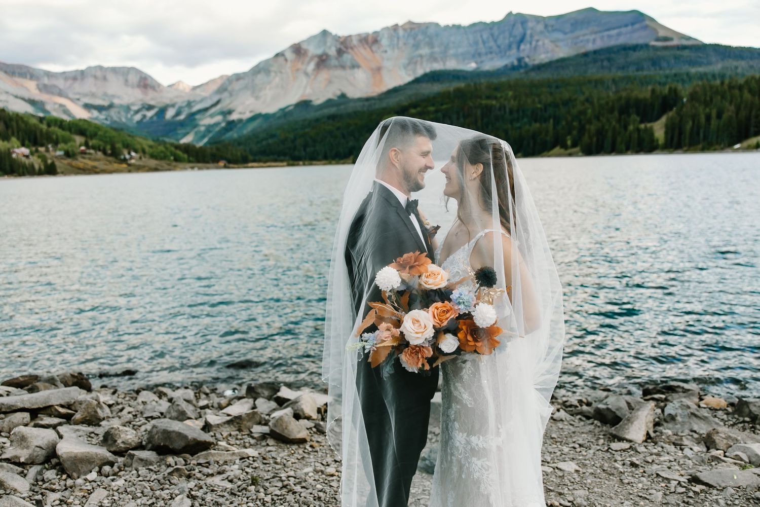 Intimate wedding portrait under flowing veil with mountain lake backdrop.