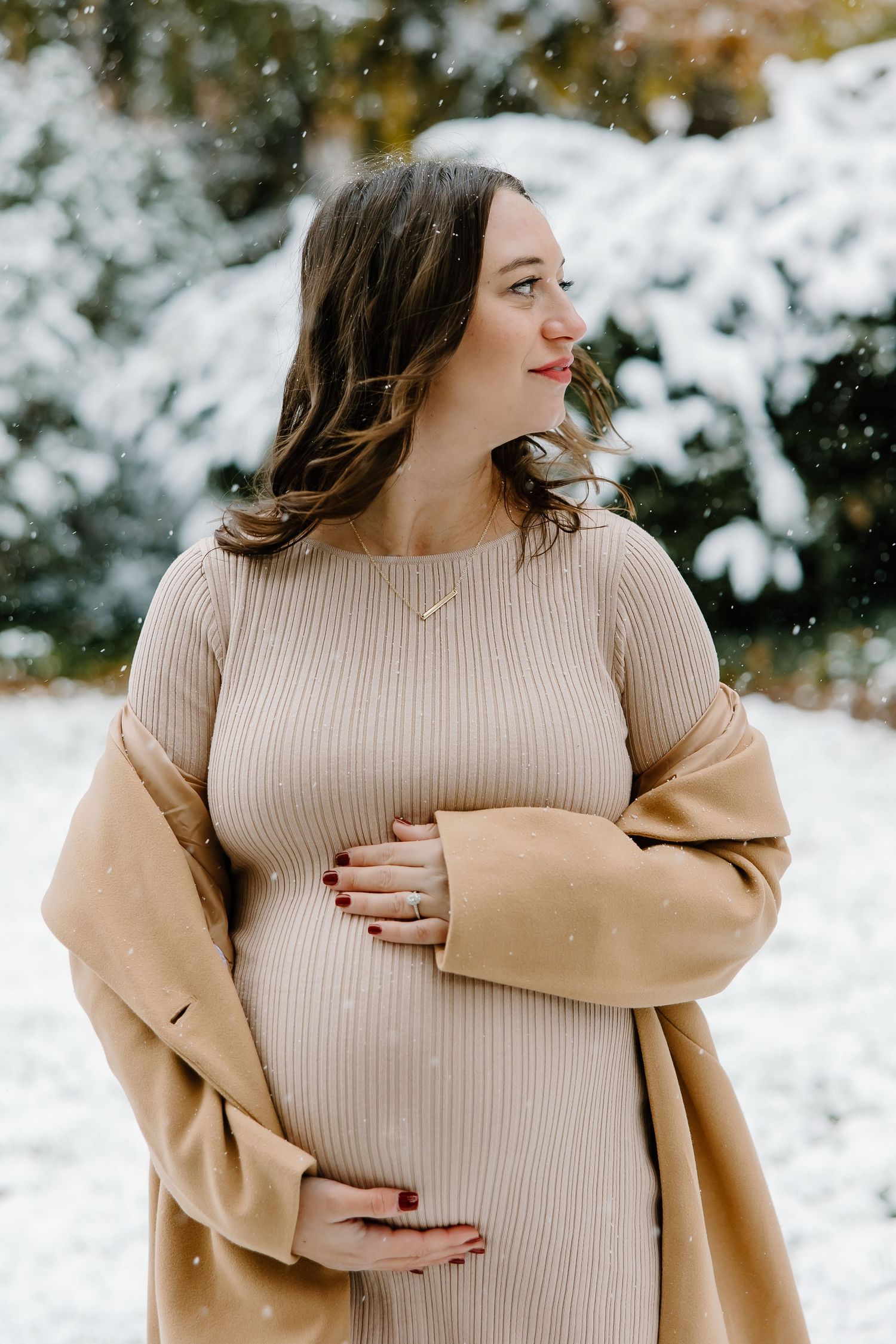 Expectant mother in cream ribbed dress and camel coat poses in snowy winter setting.