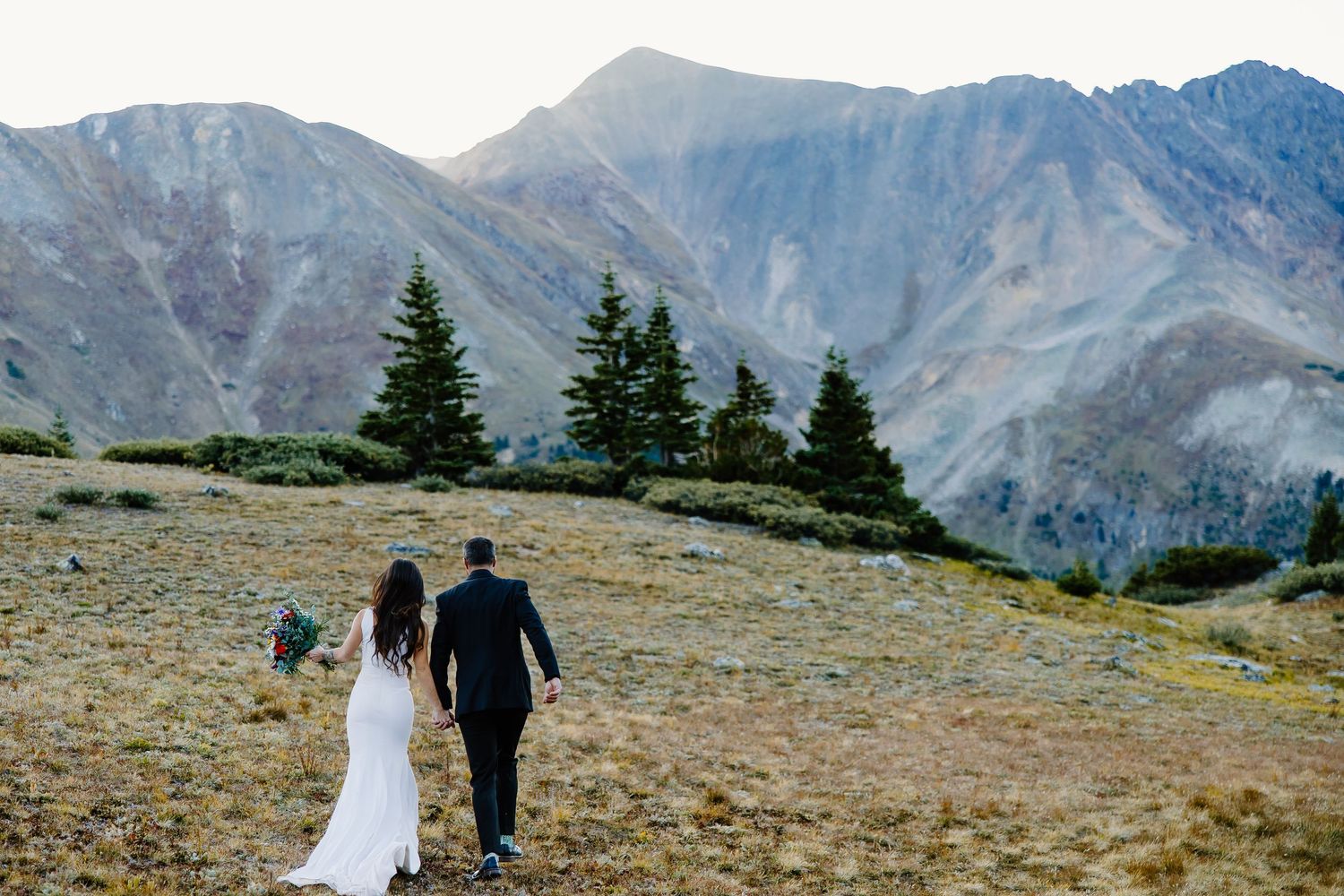 Wedding couple walks with dog along mountain trail surrounded by majestic peaks and evergreens.
