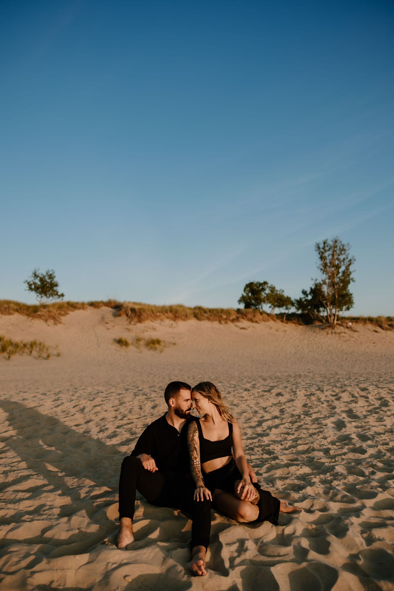 Couple sitting close together on sandy dunes during golden hour.