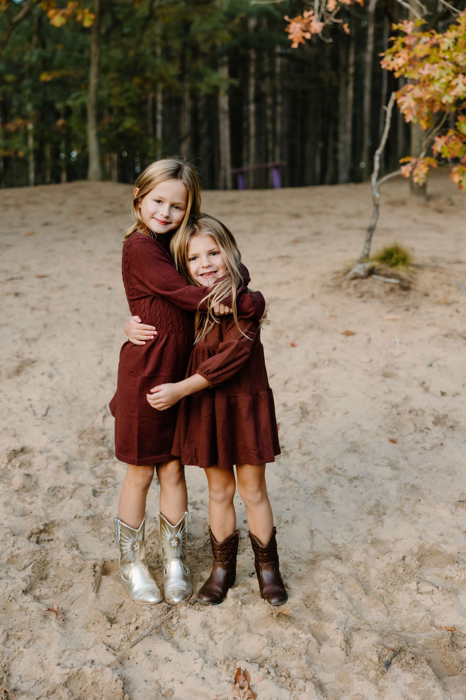 Two children in matching burgundy dresses sit together on sandy slope during autumn.