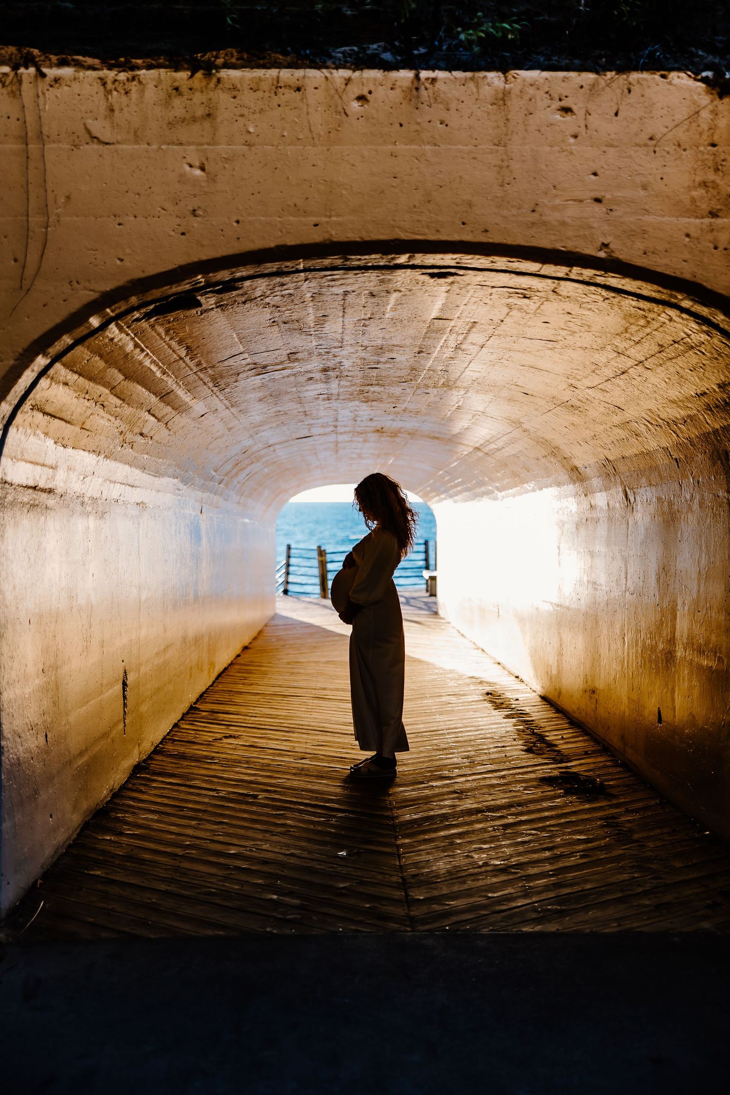 Silhouette of pregnant woman standing in sunlit wooden tunnel.