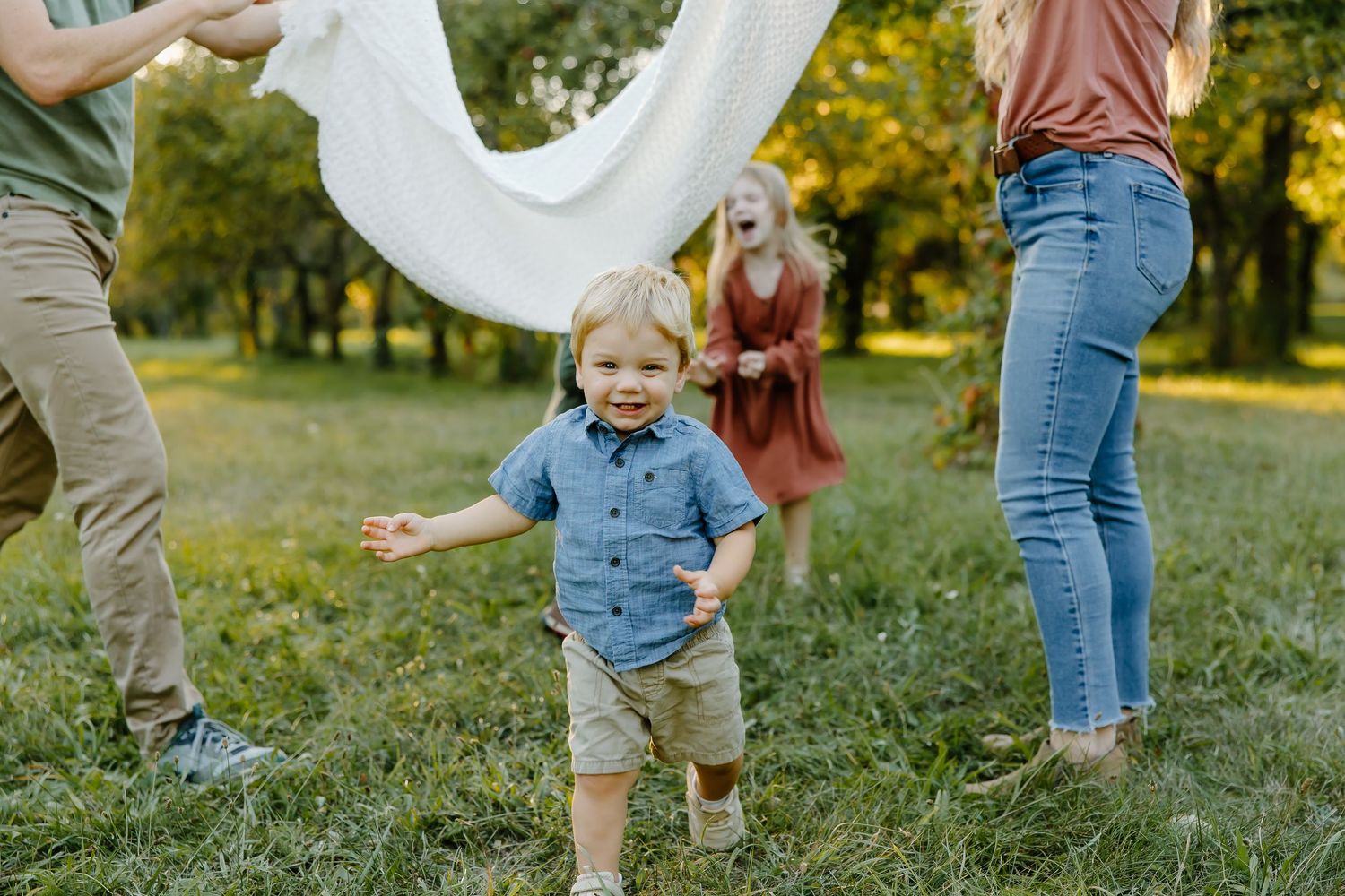 People playing with a white sheet in an orchard as a child runs underneath.