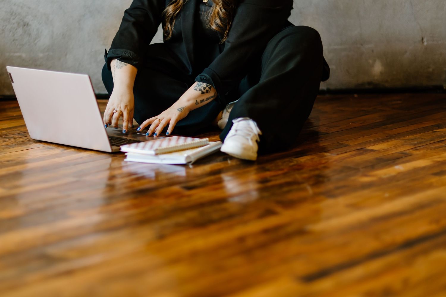 Person sitting on hardwood floor working on laptop with papers and wearing casual black clothing.