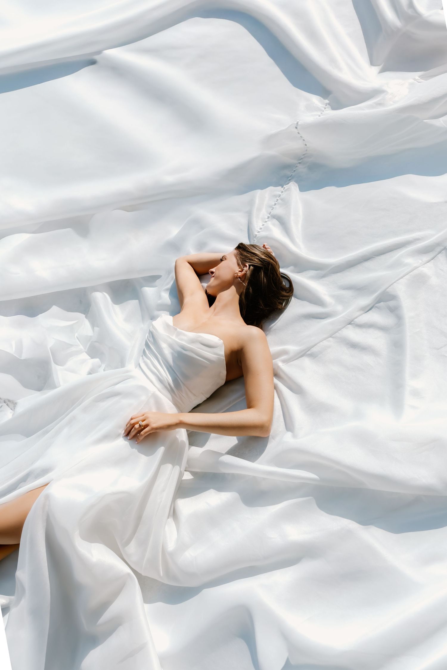 A woman in a white dress lies gracefully on flowing white fabric, creating an ethereal and elegant composition.