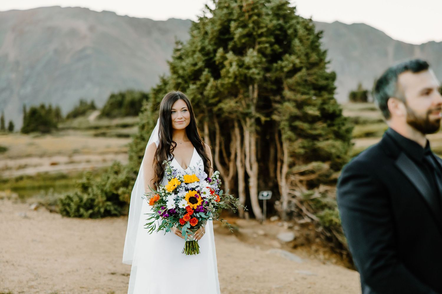 Wedding couple poses against dramatic mountain landscape with colorful bouquet.