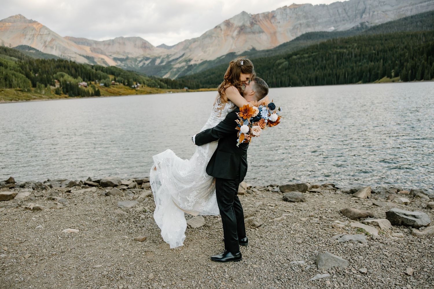 Couple sharing an intimate moment by a pristine mountain lake with dramatic peaks in the background.