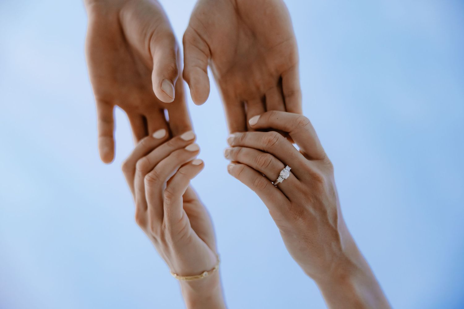 Close-up of hands with engagement rings touching against blue sky.