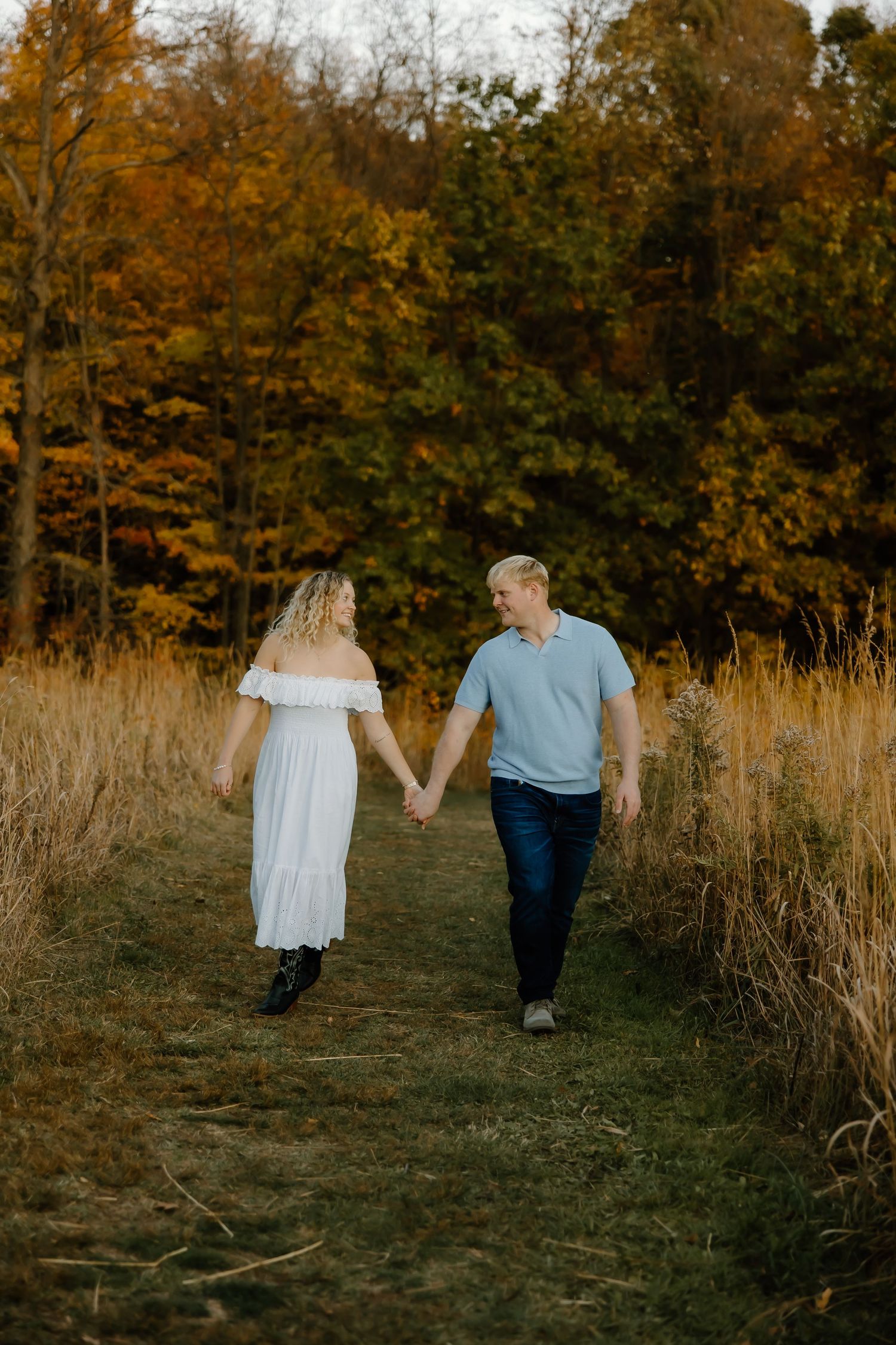 A couple walks hand in hand through a golden autumn field with trees in the background during sunset.