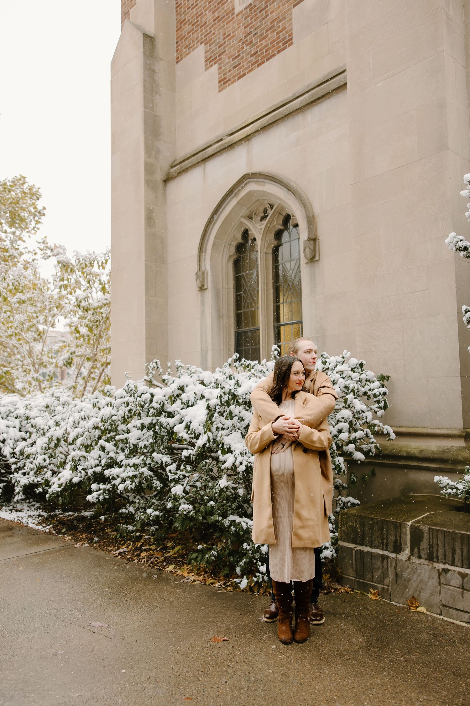 Couple embracing outside gothic church building with snow-covered bushes.
