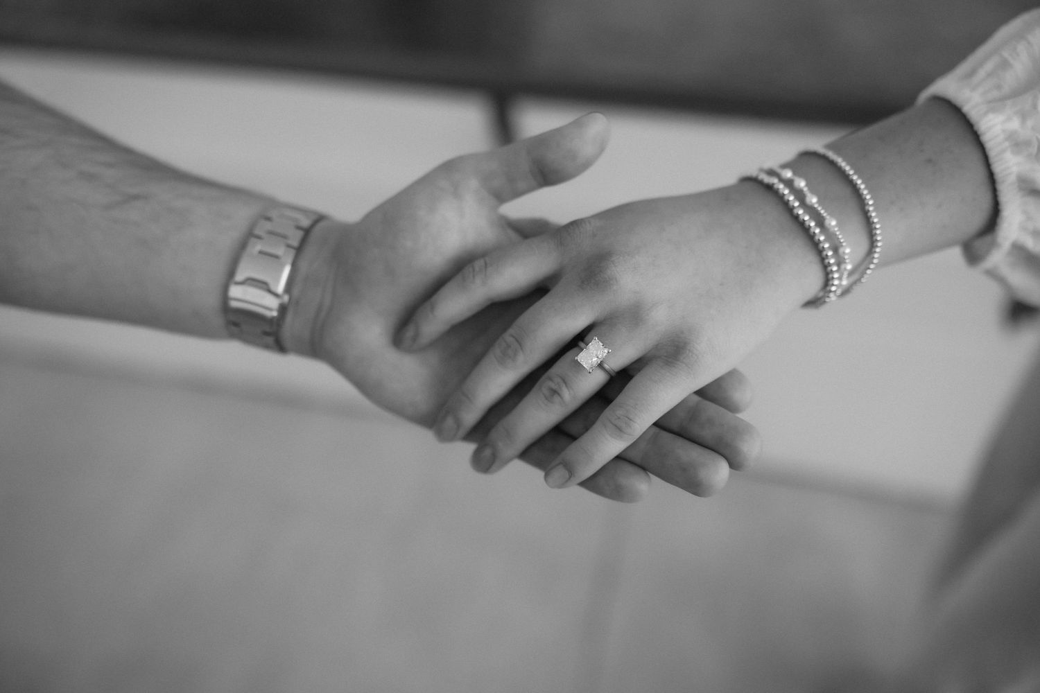Black and white photo of a couple holding hands, showing a wedding ring and bracelet, symbolizing love and commitment.