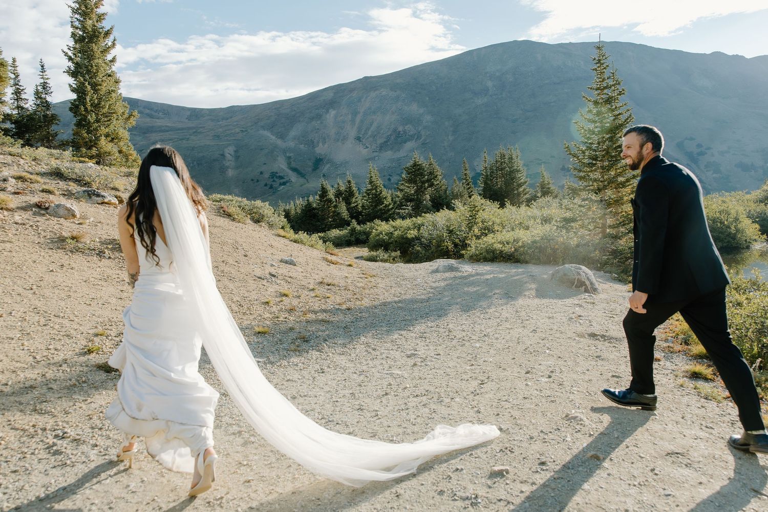 A flowing white wedding dress trails behind as the couple walks along a rocky mountain path.
