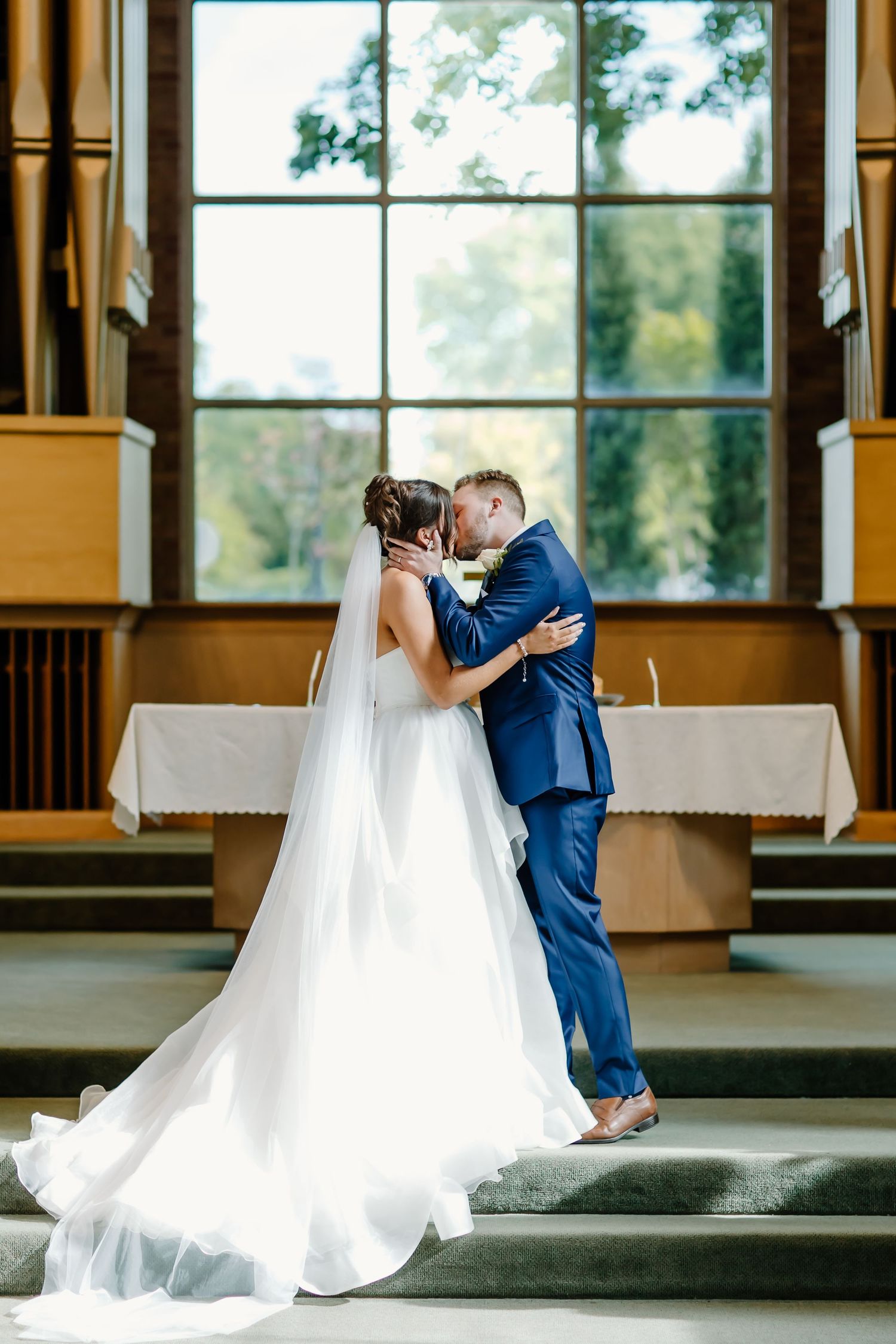 A romantic first kiss moment between newlyweds in front of a large church window.