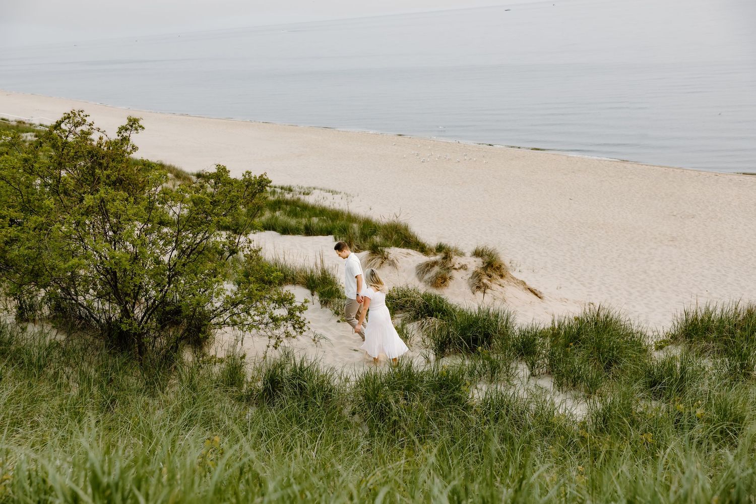 Person in white dress walking down grassy sand dunes near beach.