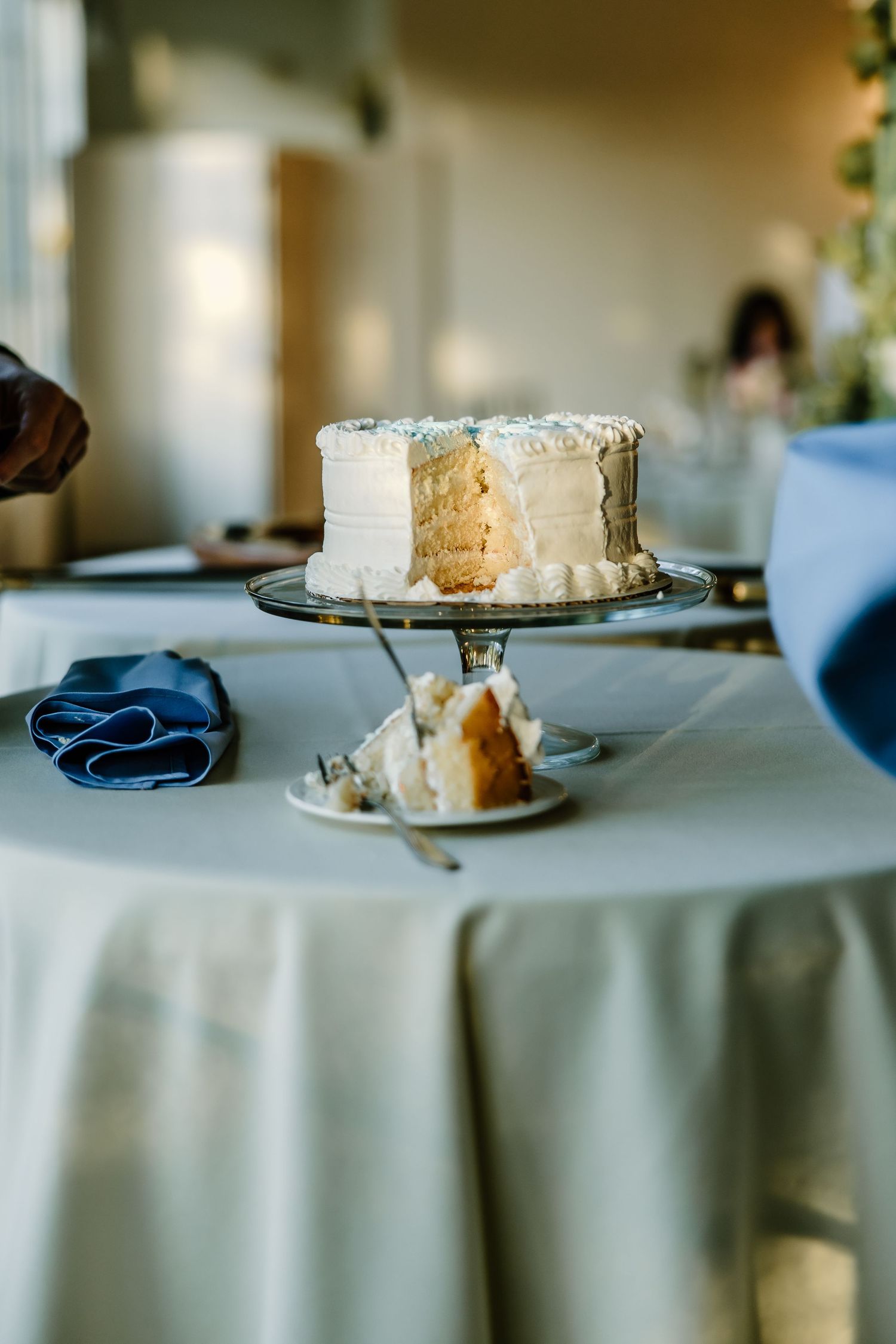 White wedding cake on silver stand with navy blue accents on table.