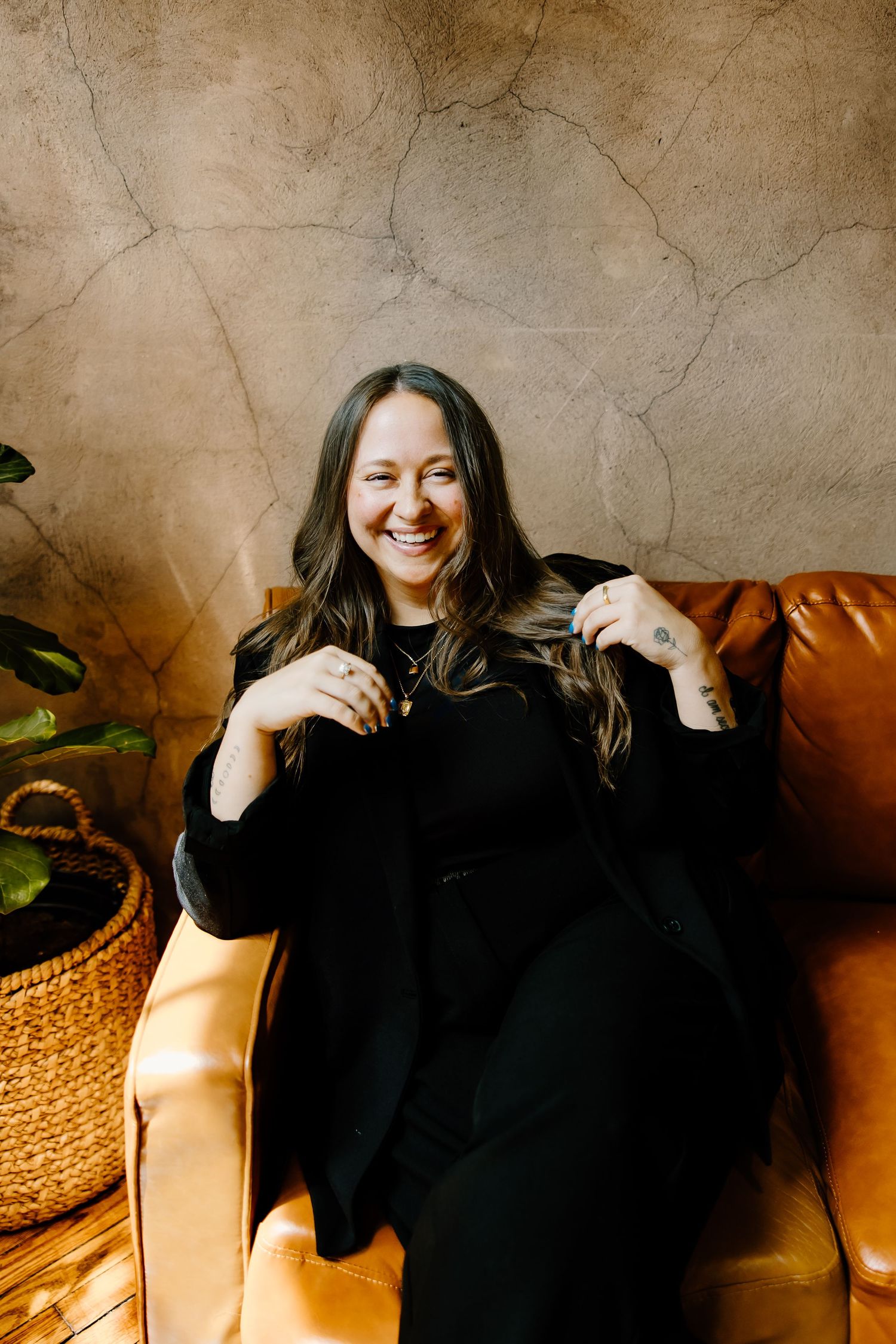 Relaxed portrait of person in black outfit sitting on tan leather couch with decorative basket nearby.