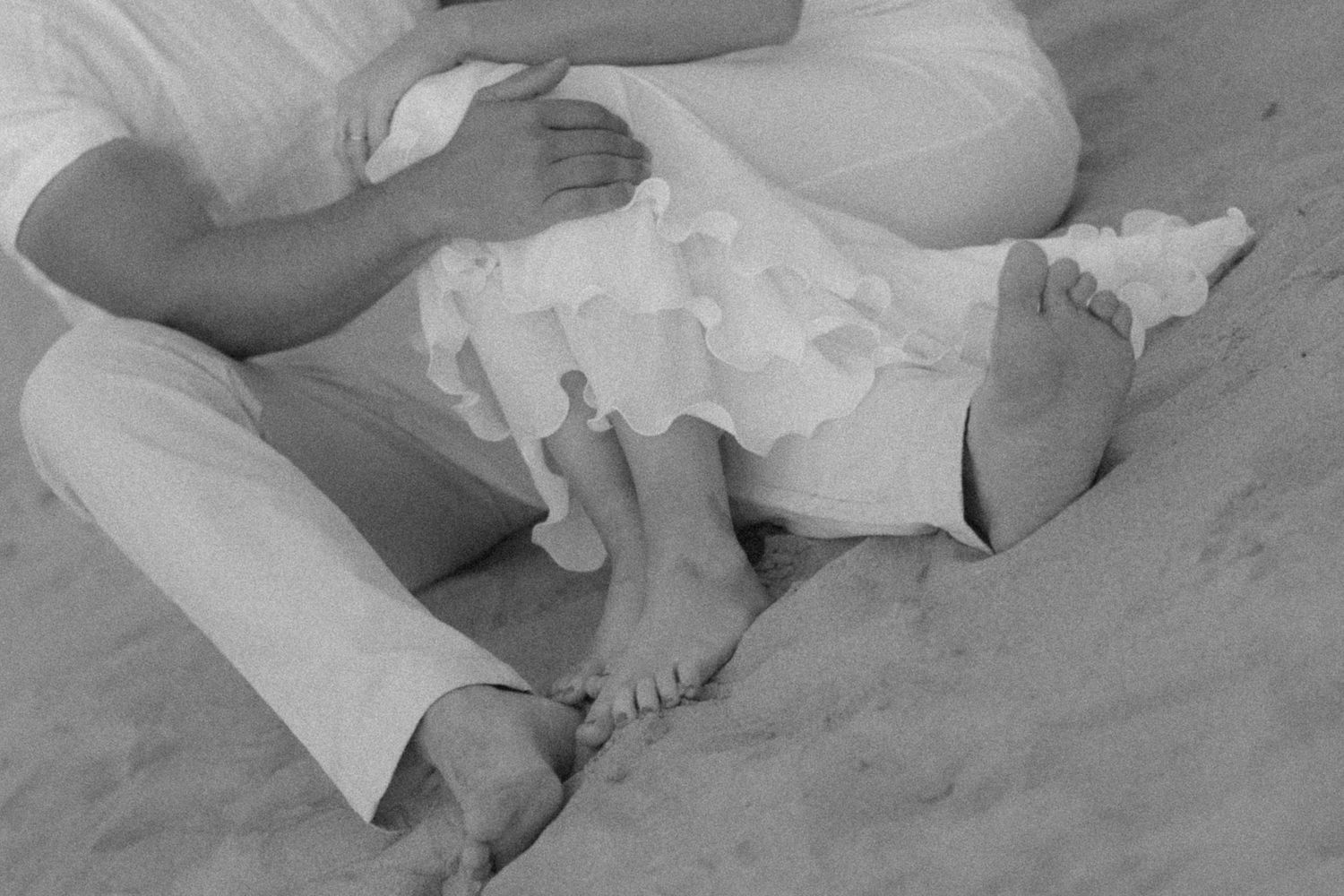 Couple sitting barefoot on sandy beach, black and white photo showing legs and white dress.