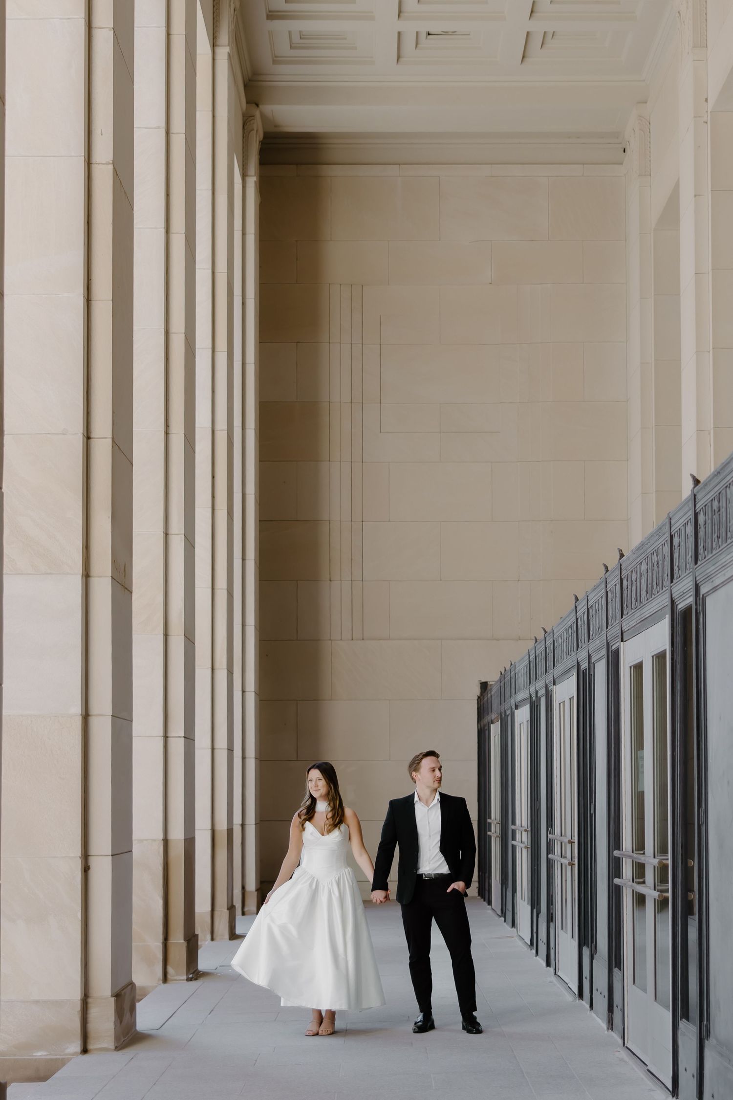 Bride and groom walk hand in hand along a grand colonnade with tall stone pillars at their wedding venue.
