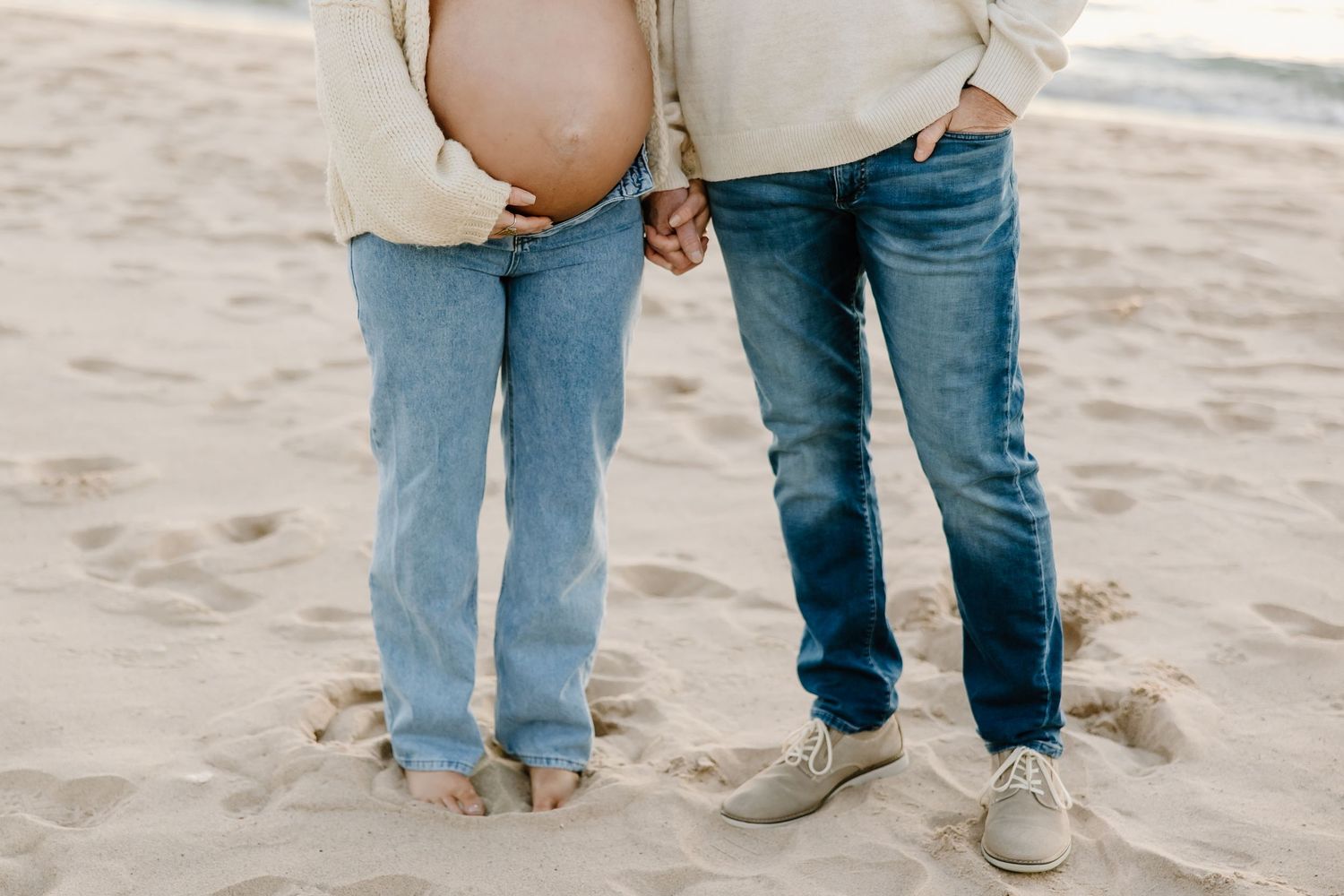 Close-up of feet and legs in jeans standing on a sandy beach.