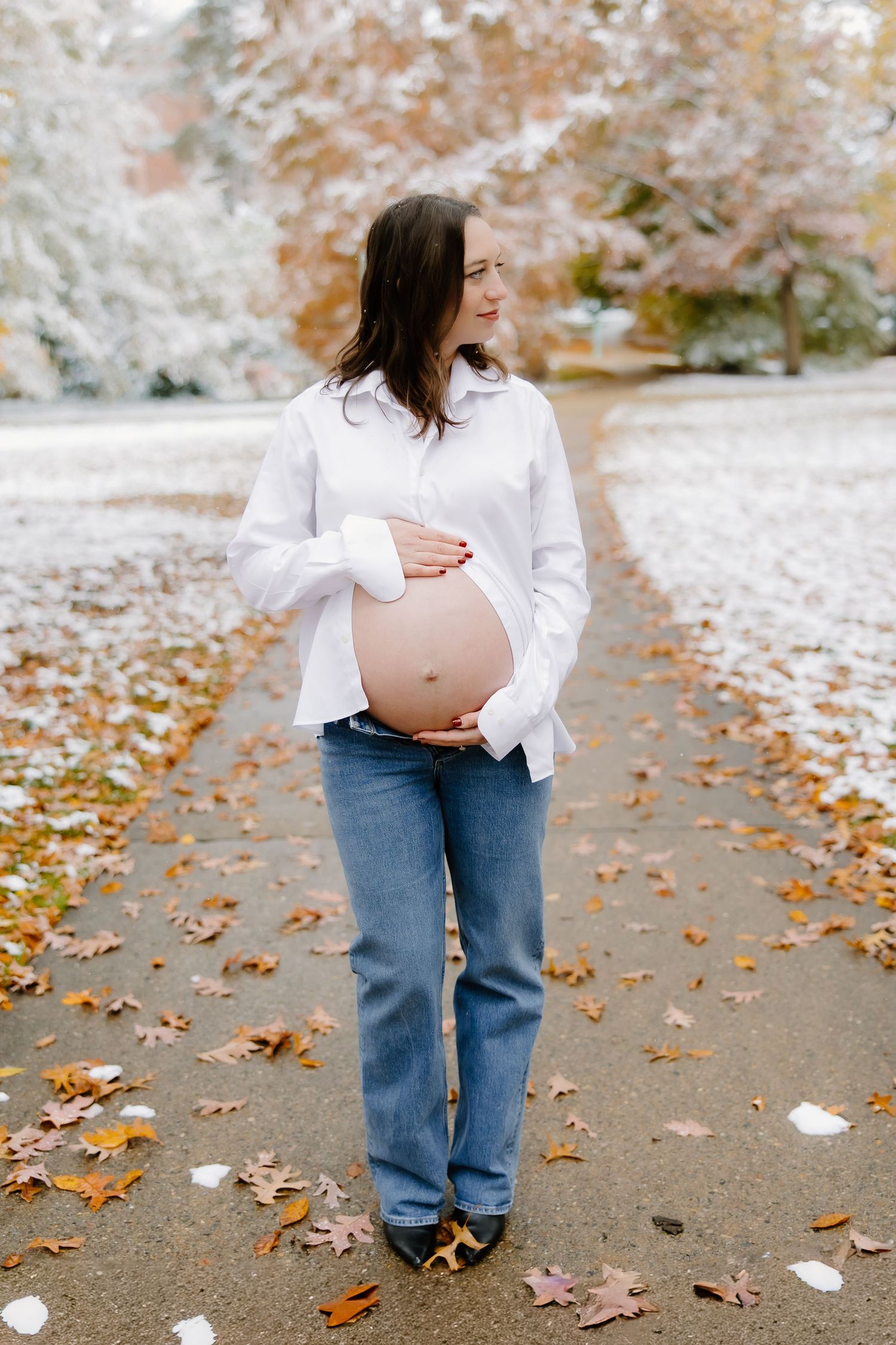 Pregnant woman in white shirt and jeans standing on snowy path with fall leaves.