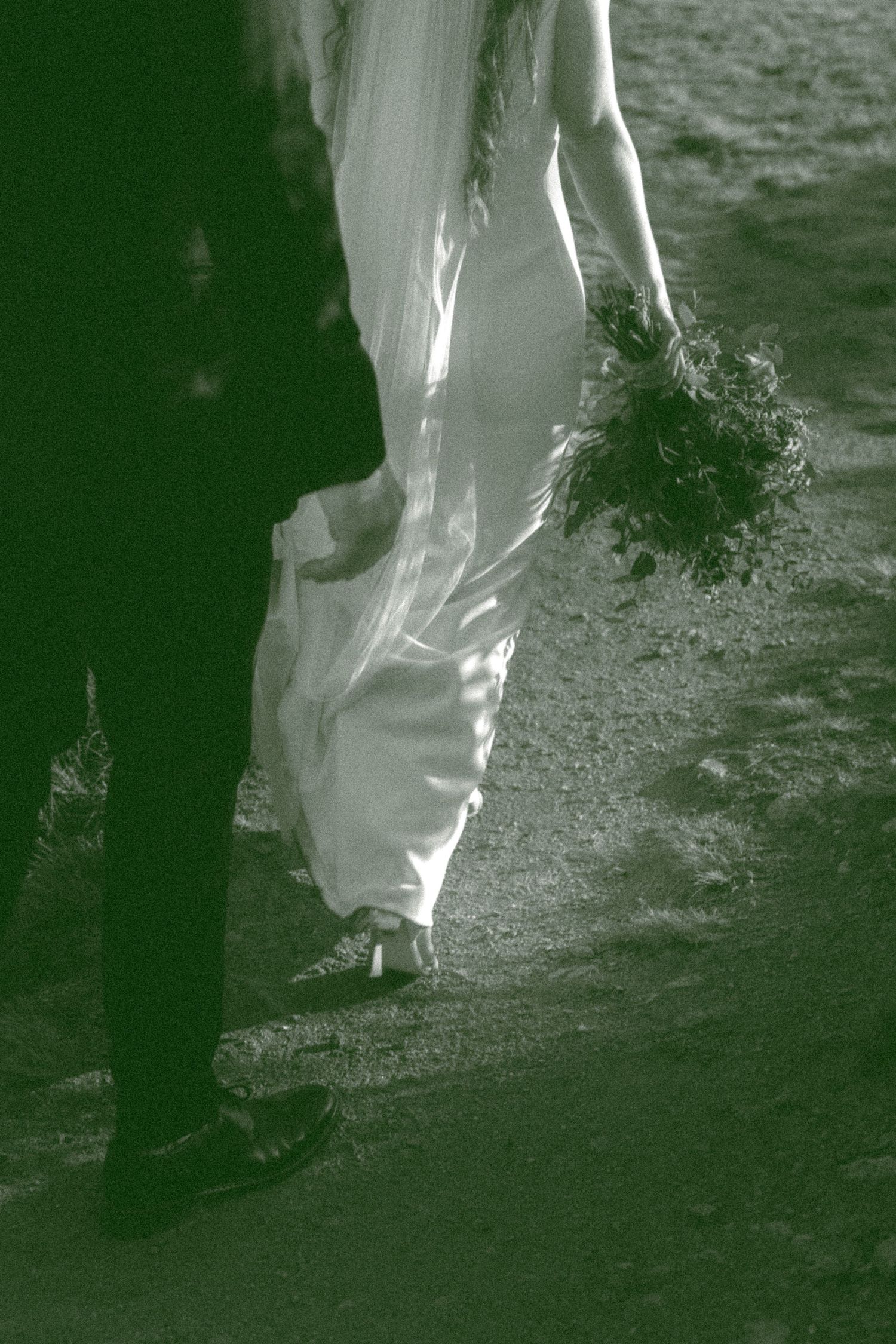 Close-up of wedding dress details as couple walks along a rocky path at dusk.