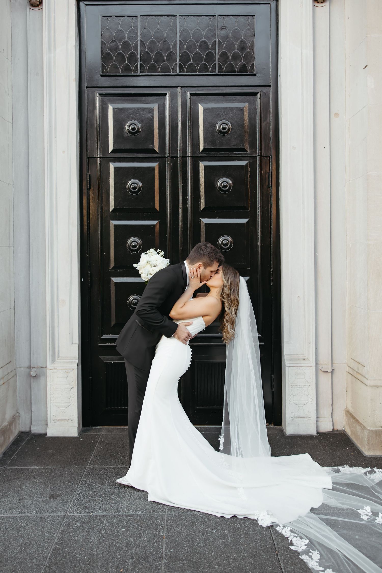 Dramatic wedding portraits in front of ornate black doors with flowing white veil.