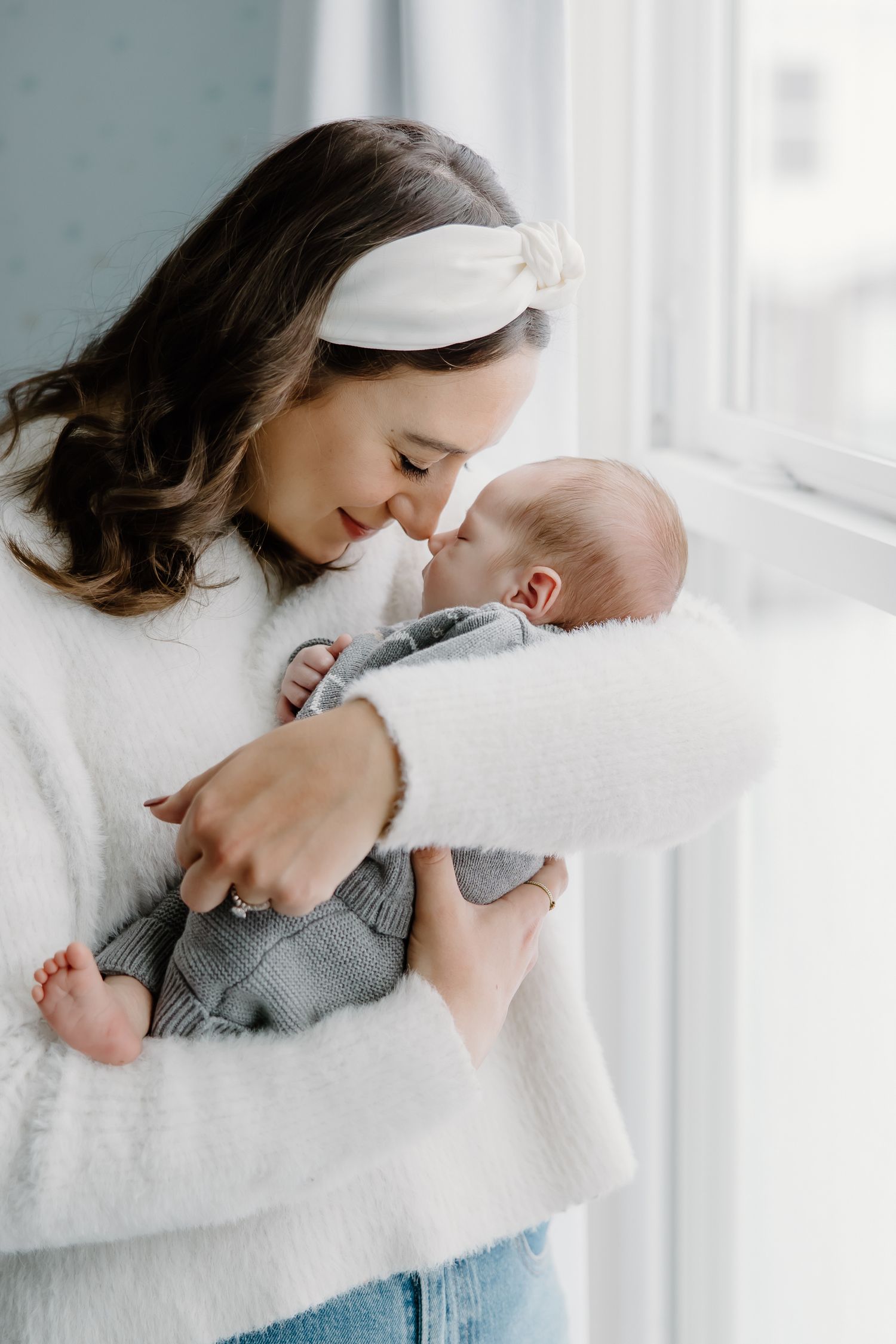 Sweet candid moment between parent and baby in white sweater by sunlit window.