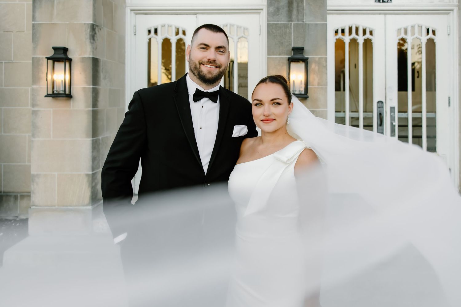 Elegant wedding couple posing in front of white stone building with classic lanterns.