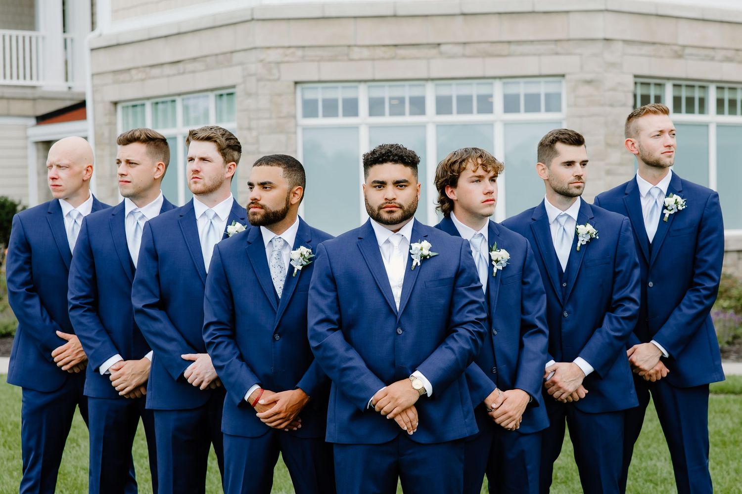 Groomsmen in matching navy suits stand in a line outdoors.
