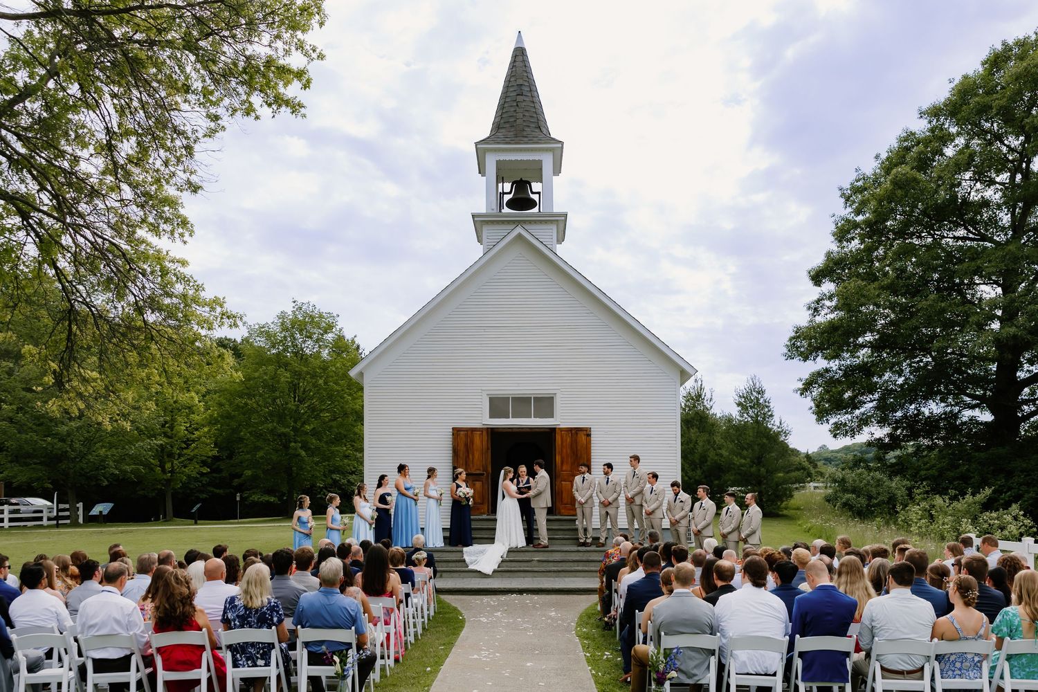 Outdoor wedding ceremony at a white country chapel with bridal party and seated guests on a sunny day.