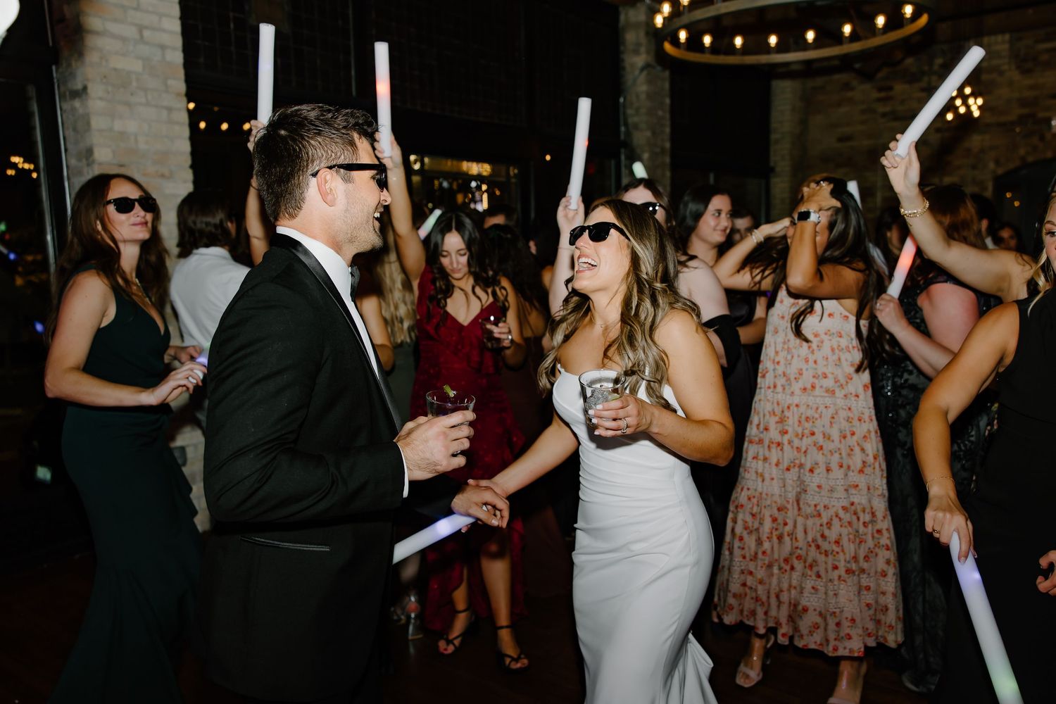 Wedding celebration with guests dancing under warm string lights and brick walls.
