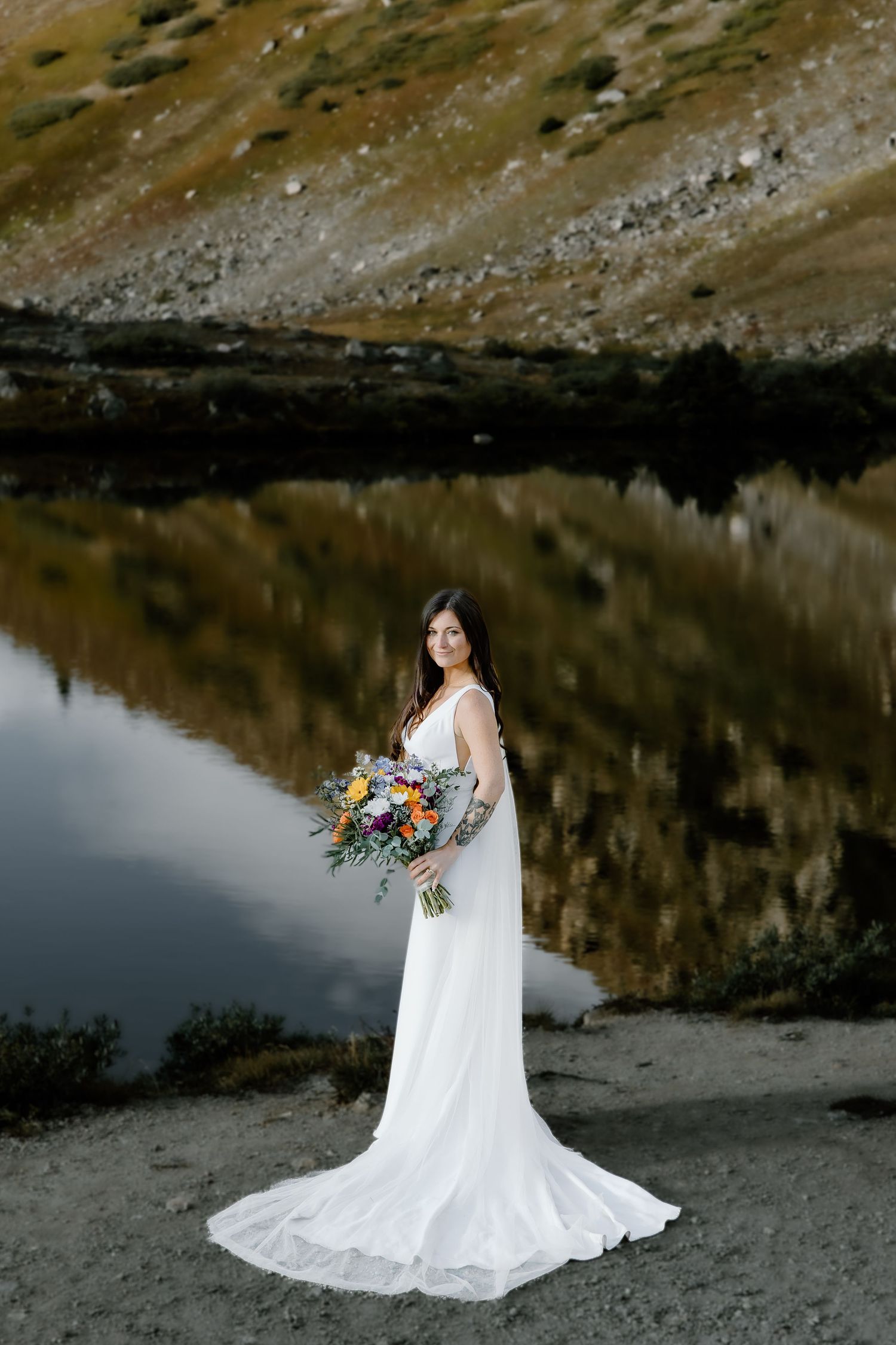 Bride in flowing white dress poses by mountain lake with dramatic reflection.